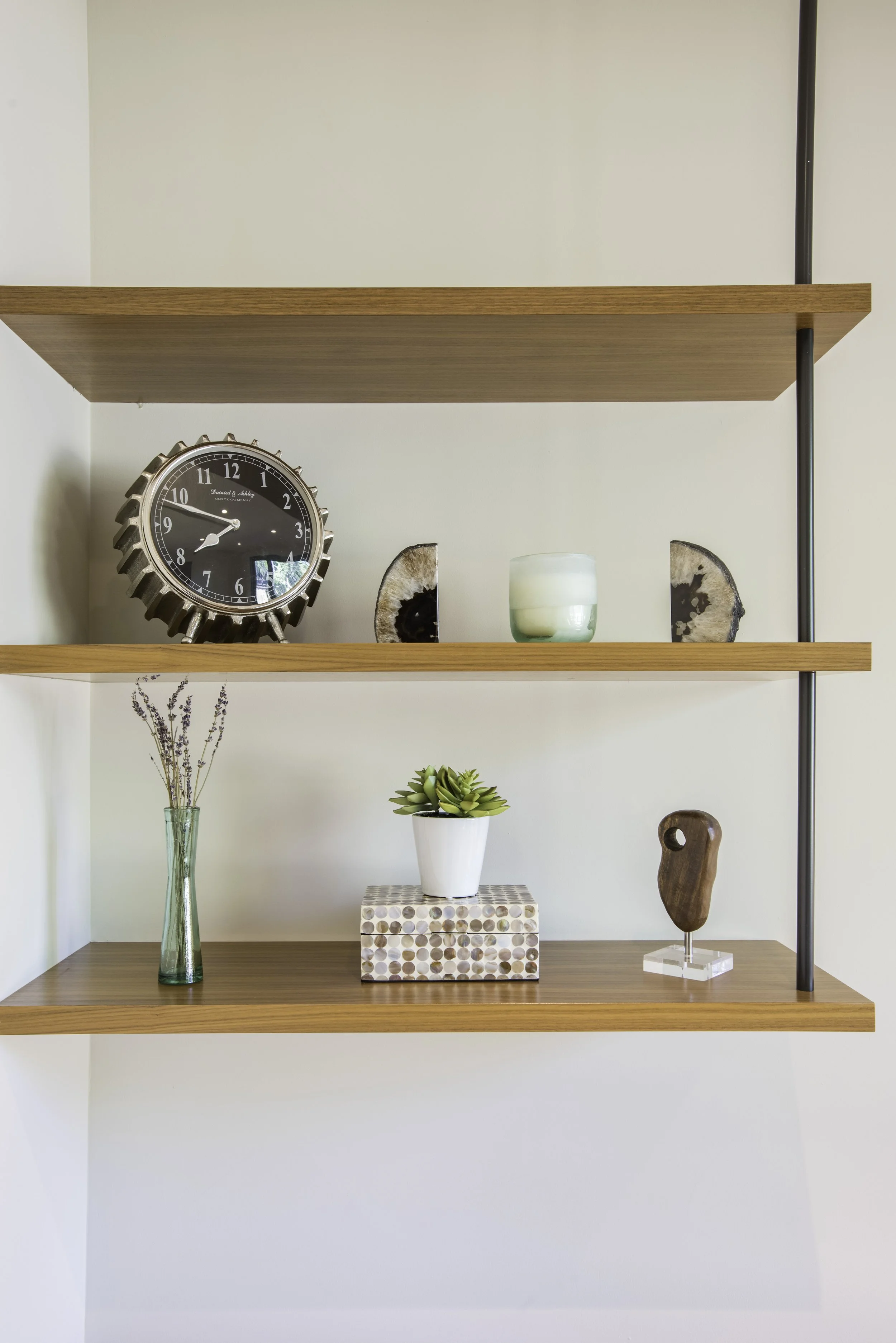 Wooden shelves with decorative items including a black clock, two rocks, a green candle holder, a vase with lavender, a potted succulent, a box, and a wooden sculpture.