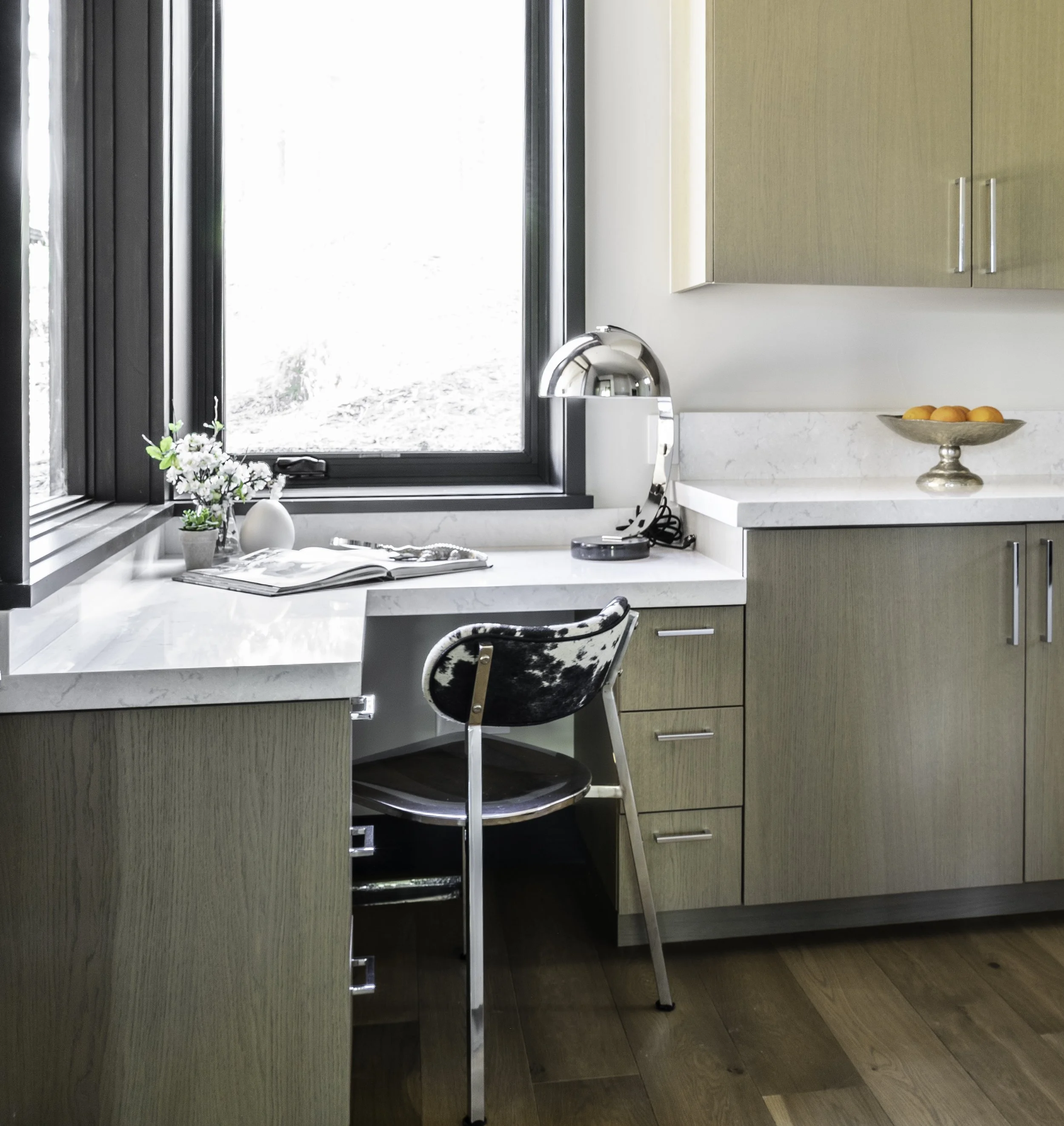 A kitchen corner with a large window, a marble countertop, a black and silver chair, a small table lamp, a vase with flowers, a book, and a dish with oranges.