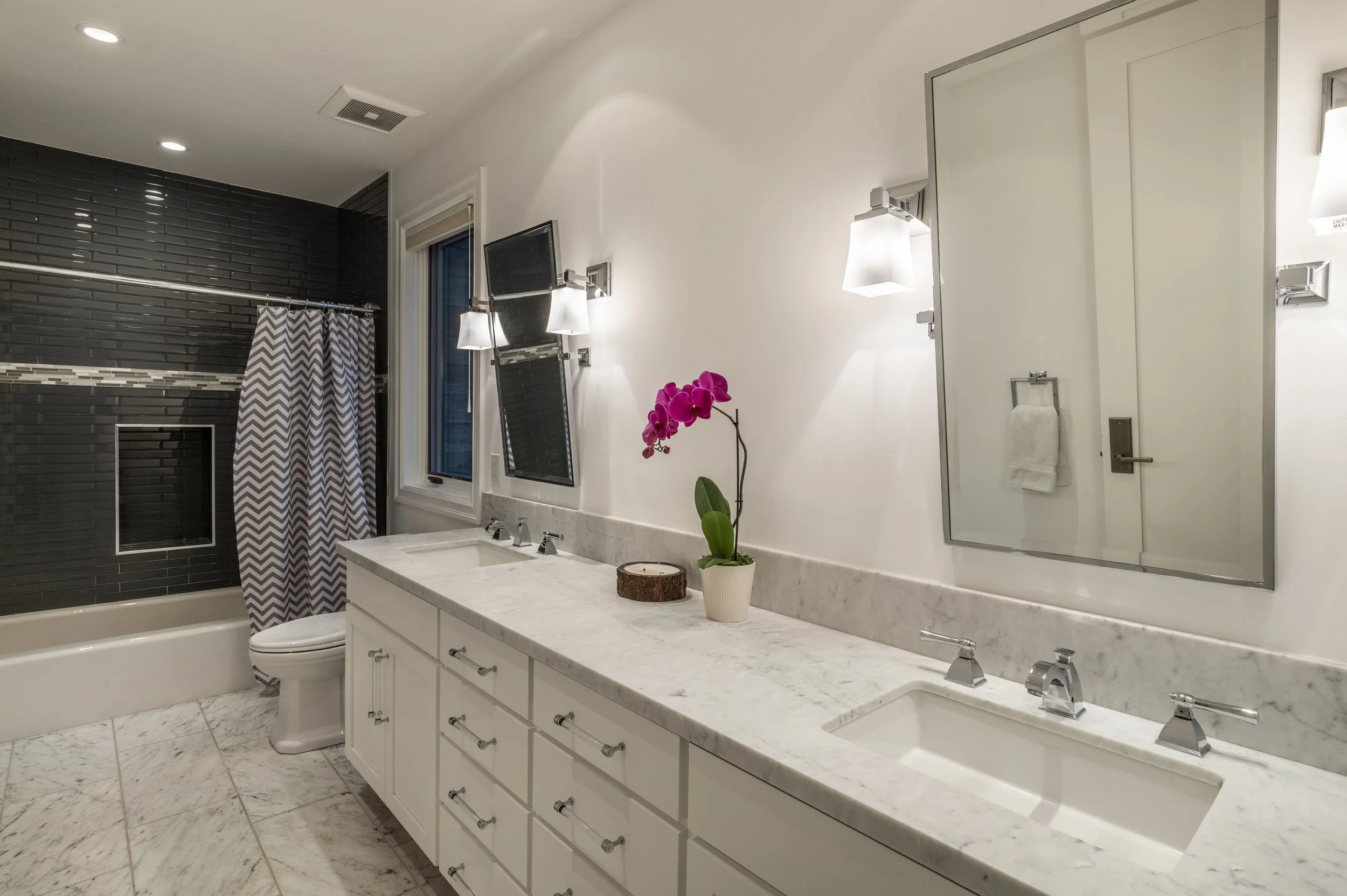 Modern bathroom with a white marble countertop, dual sinks, a large mirror, white cabinets, a pink orchid plant, a black tiled shower area with a chevron-patterned curtain, and black wall tiles with small mosaic accents.