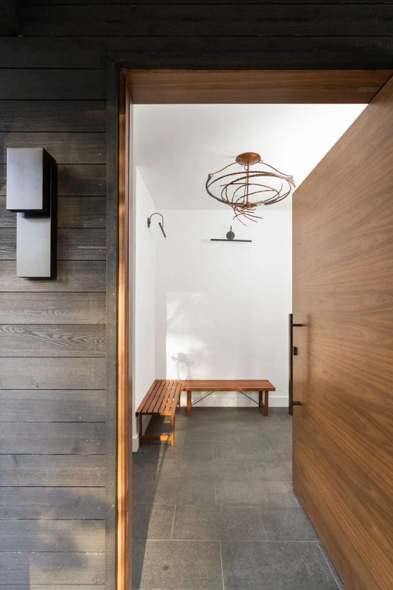 View of a modern hallway with a wooden bench and a minimalist chandelier hanging from the ceiling, seen through a partially open wooden door.