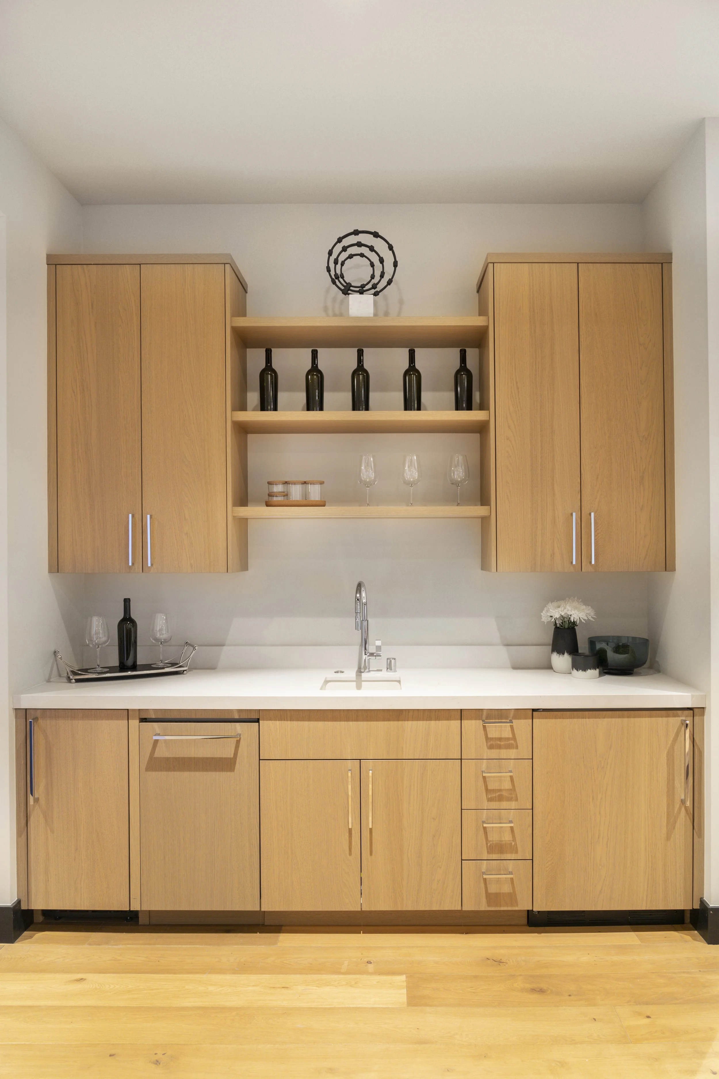 A minimalist kitchen with light wooden cabinets, a white countertop, and a modern faucet. Open shelves display wine bottles and glasses, with decorative items including a vase with white flowers and a dark bowl.