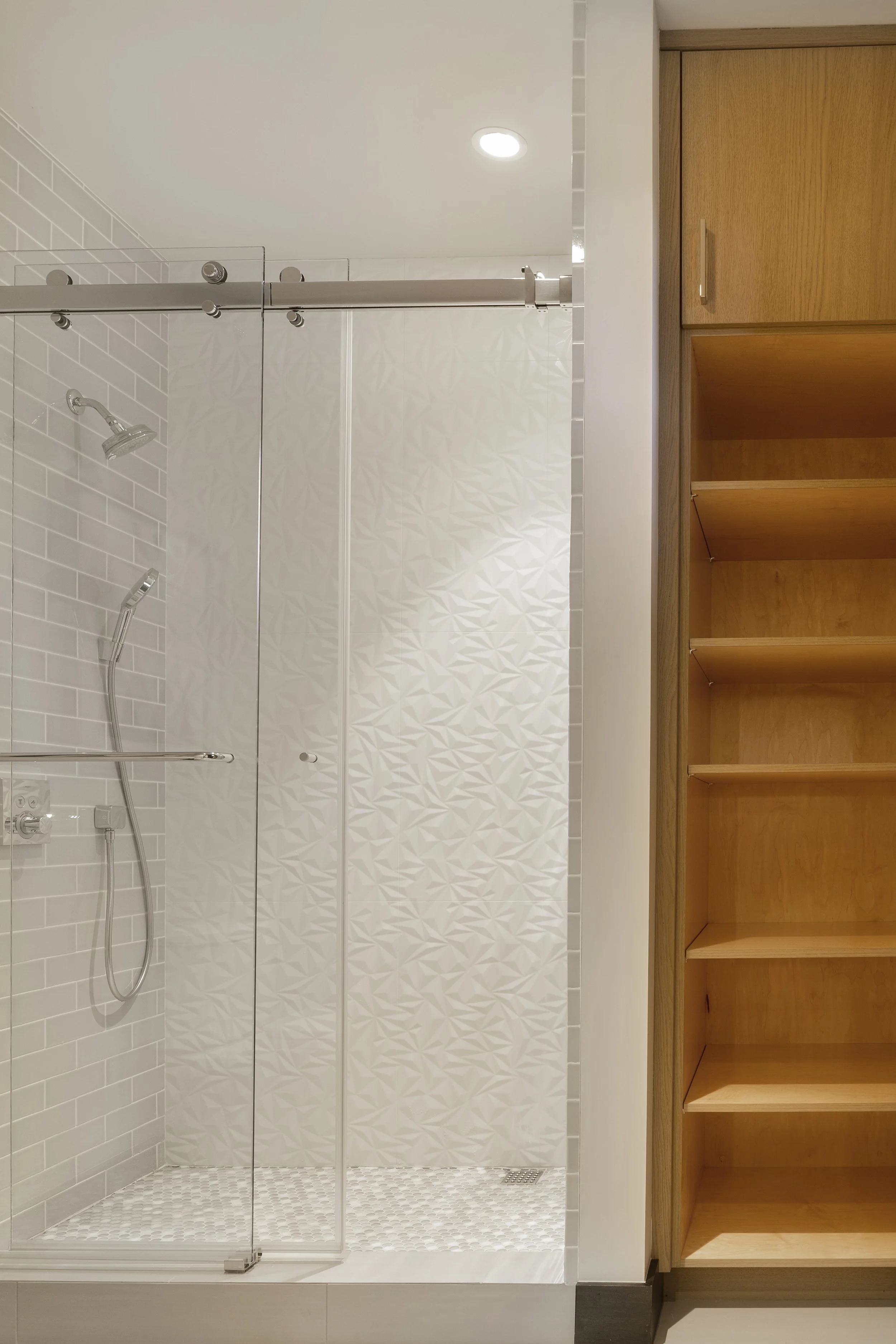 Modern walk-in shower with white patterned tiles, glass doors, and a wooden shelving unit nearby.
