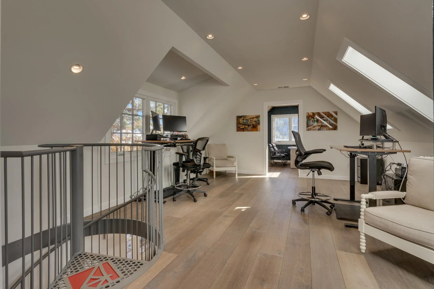 Attic converted into a home office with three desks, chairs, computers, and two paintings on the walls. Skylights and a window illuminate the space, which has light wood flooring and white walls.