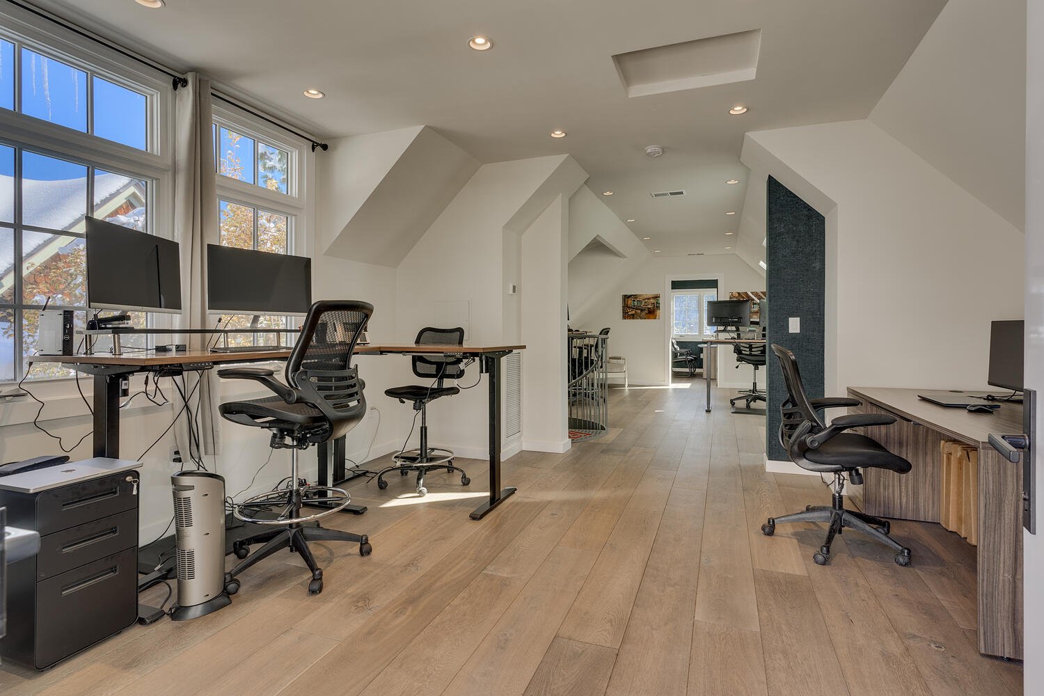 Home office with multiple desks, black office chairs, dual monitors, and large windows with white curtains, in a bright, modern space with wood flooring.