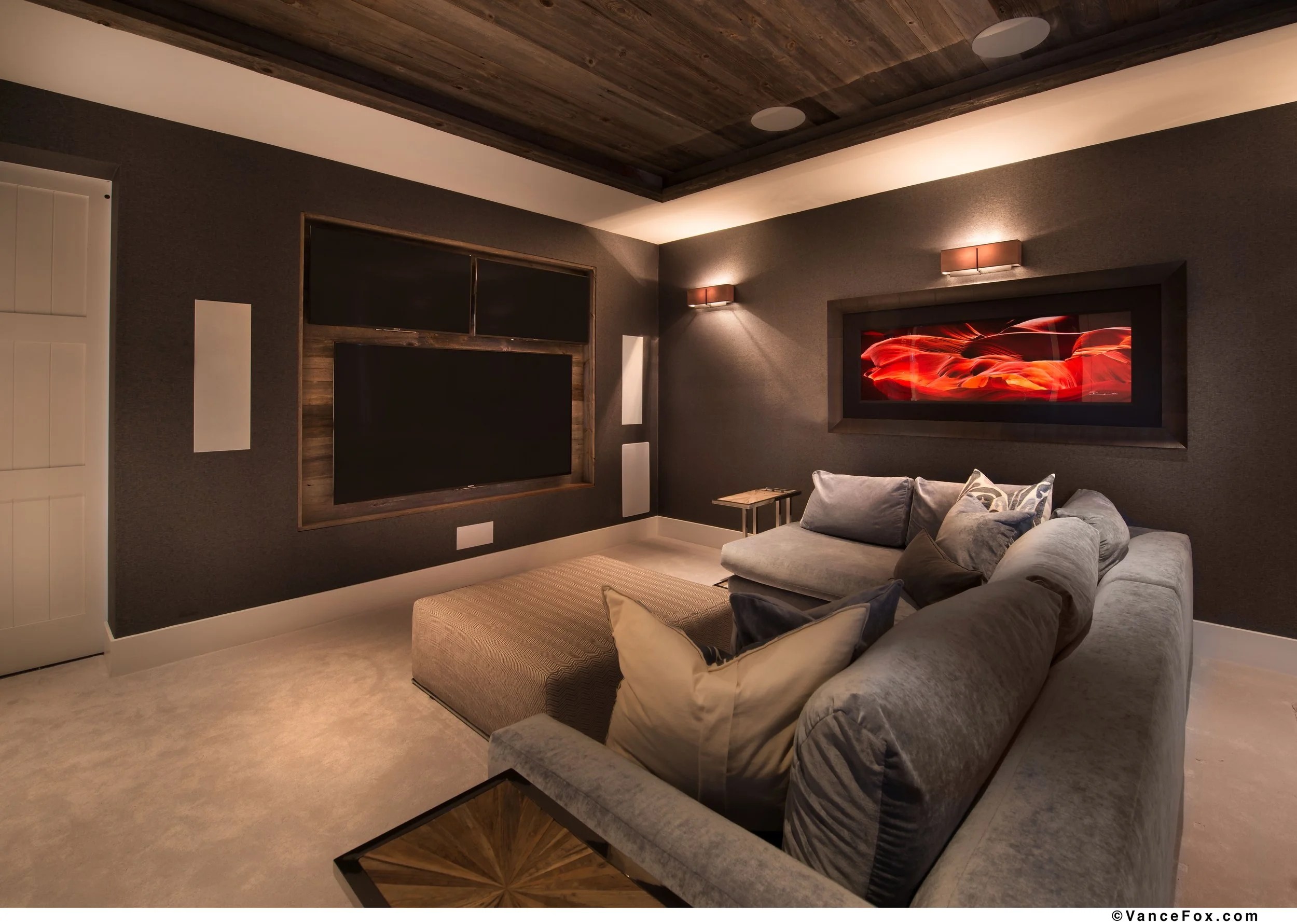 A cozy transitional media room with a large denim sectional, a rectangular ottoman, wall-mounted TVs, and contemporary artwork on textured wallpapered walls with wood ceiling, and moody lighting.