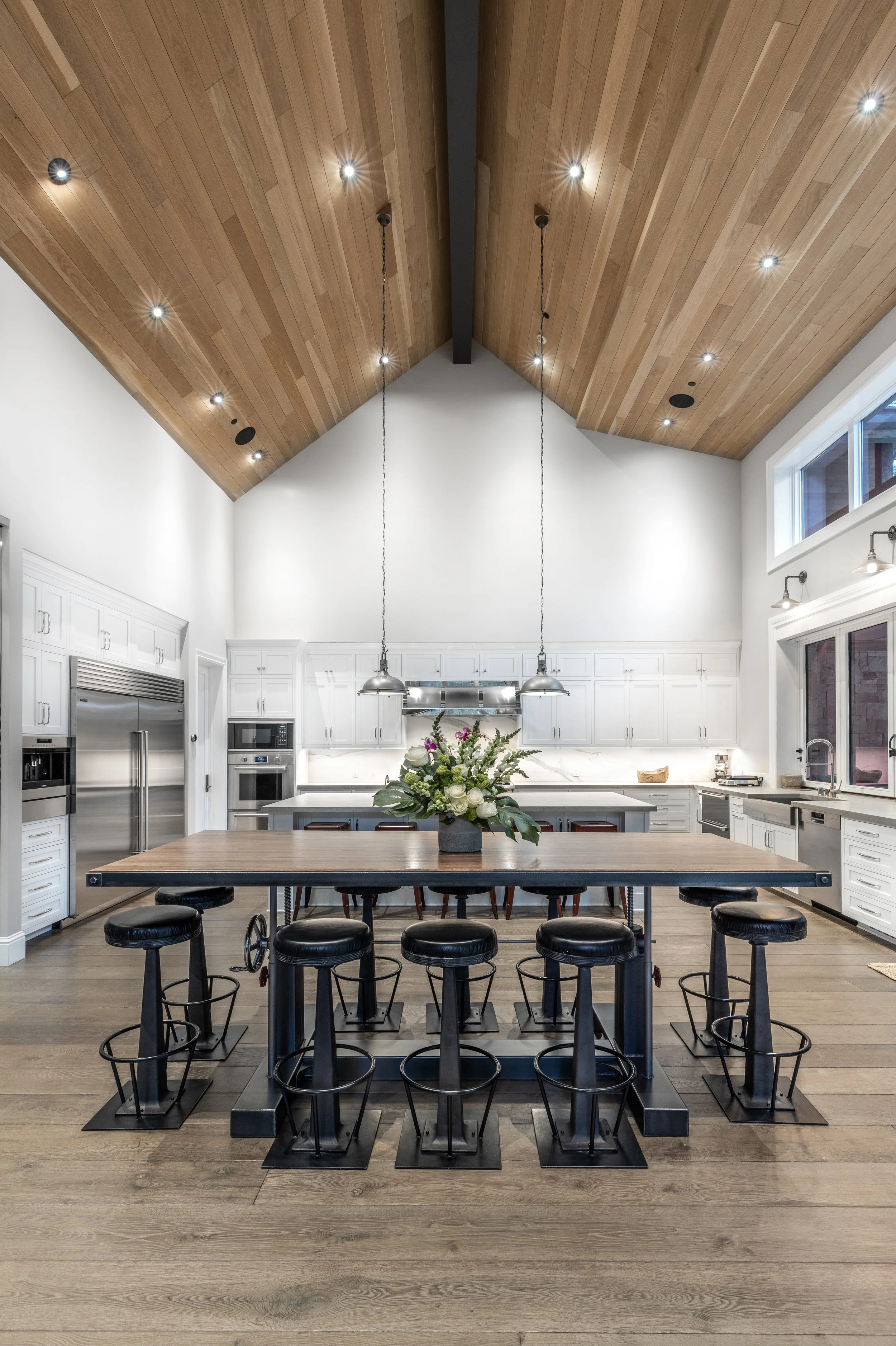 A modern kitchen with a large wooden island, black bar stools, white cabinetry, stainless steel appliances, and a vaulted wooden ceiling with recessed lighting and pendant lights.