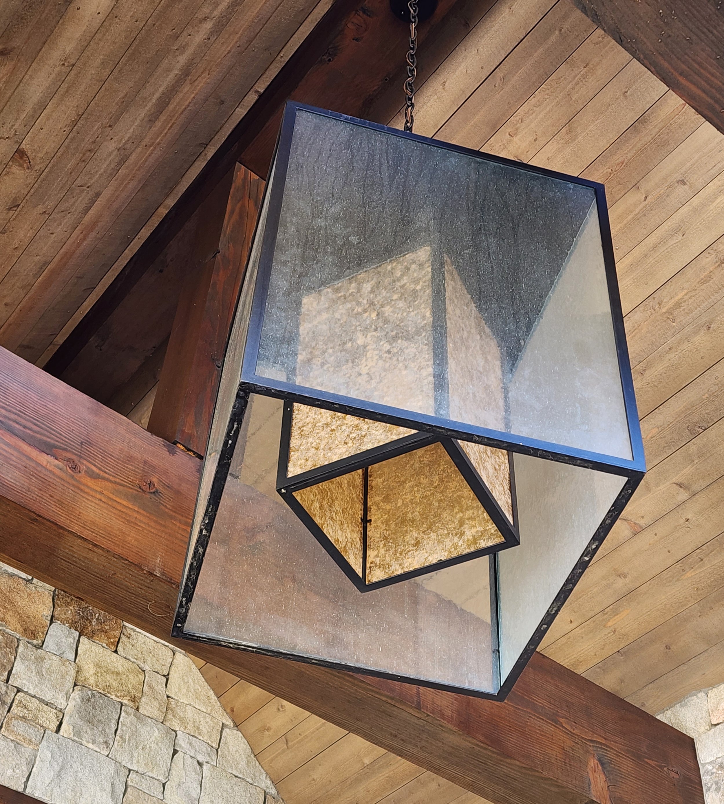 A modern geometric pendant light fixture hanging from a wooden ceiling, with a combination of black metal frame and glass panels on the outside and a mica and black framed interior shade. Custom designed by Emily Roose.