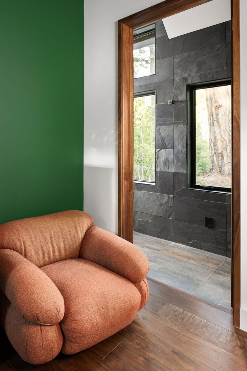 A cozy room corner with a peach-colored plush armchair, a green wall, and a doorway with a wooden frame leading to a tiled area with gray stone walls and windows showing outdoor trees.
