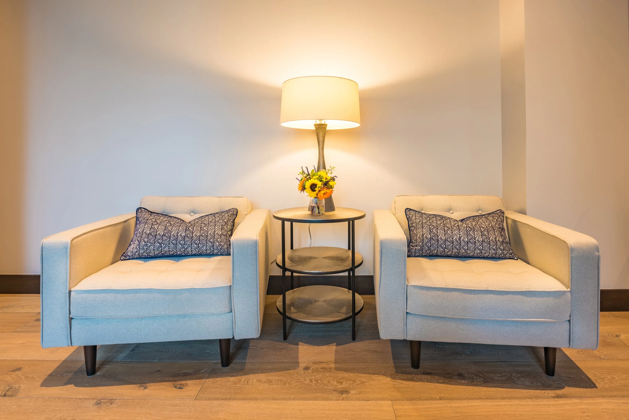 Two beige upholstered armchairs with dark wooden legs, each with a navy patterned rectangular pillow, flanking a round black side table with three shelves. On the table is a tall lamp with a beige shade, and a small vase of sunflowers and other yello