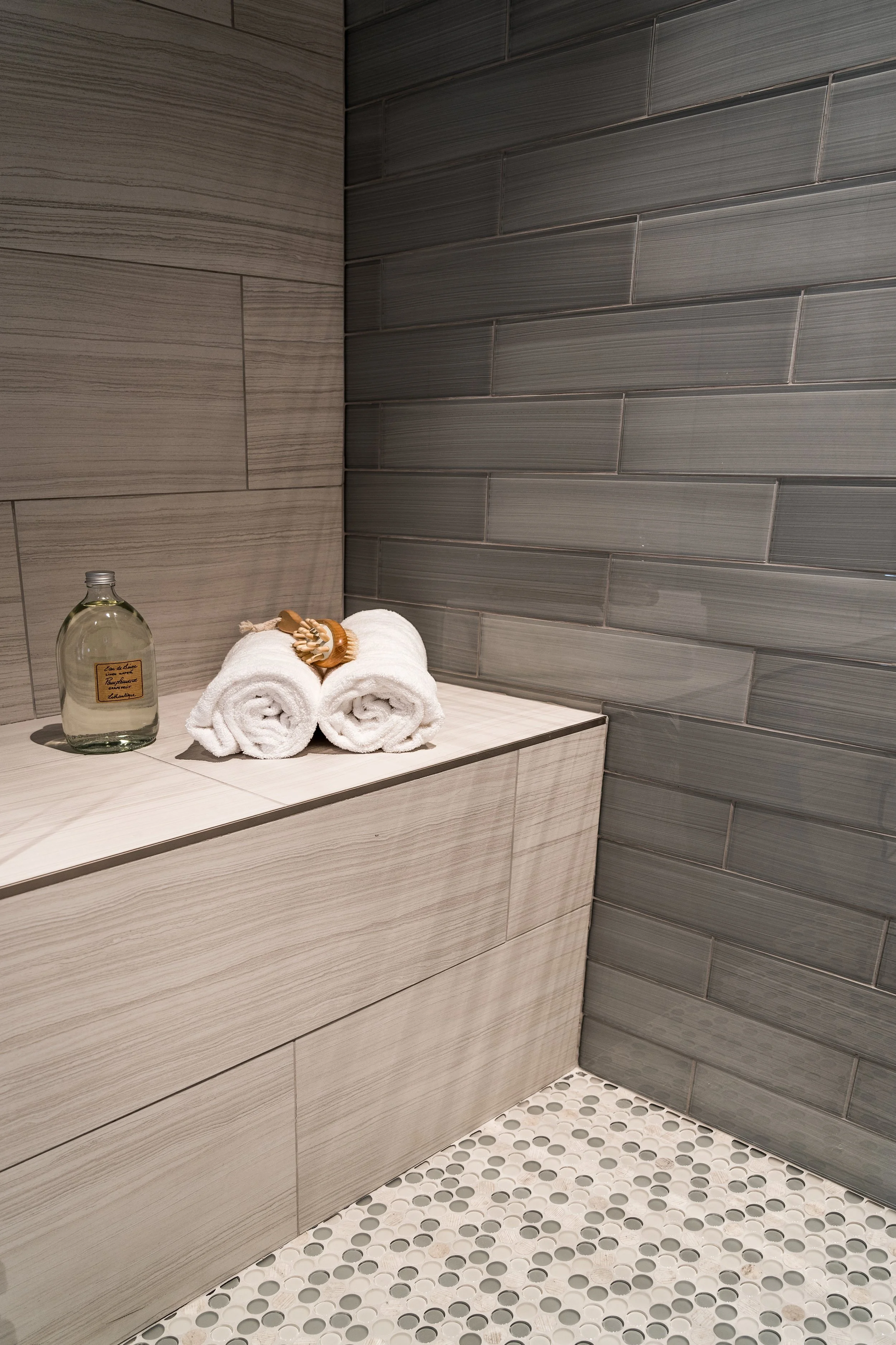 A bathroom corner featuring a wooden ledge with a glass bottle of lotion, two rolled white towels, and a wooden bath brush. The background has gray tiles and a patterned tile floor.