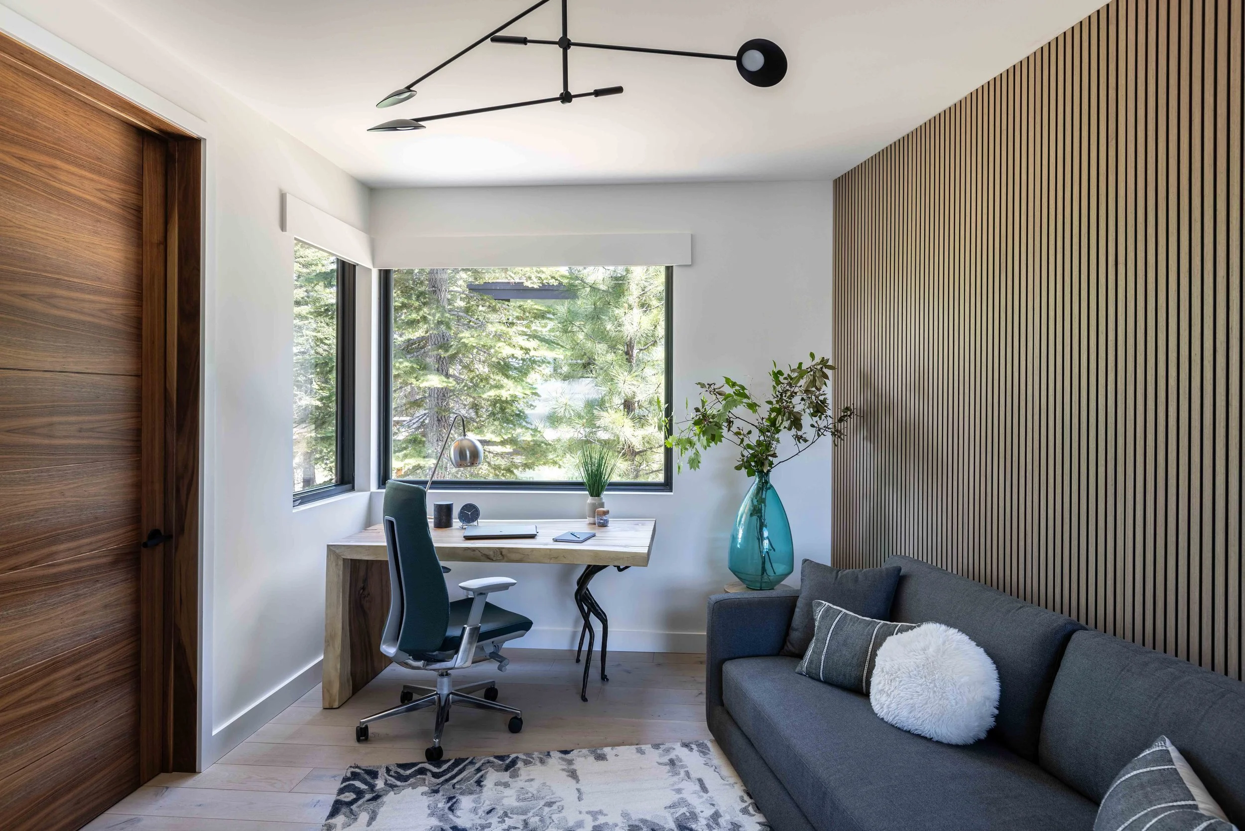 Modern home office with large window, wooden wall, gray sofa with pillows, desk with office supplies, and decorative plants.
