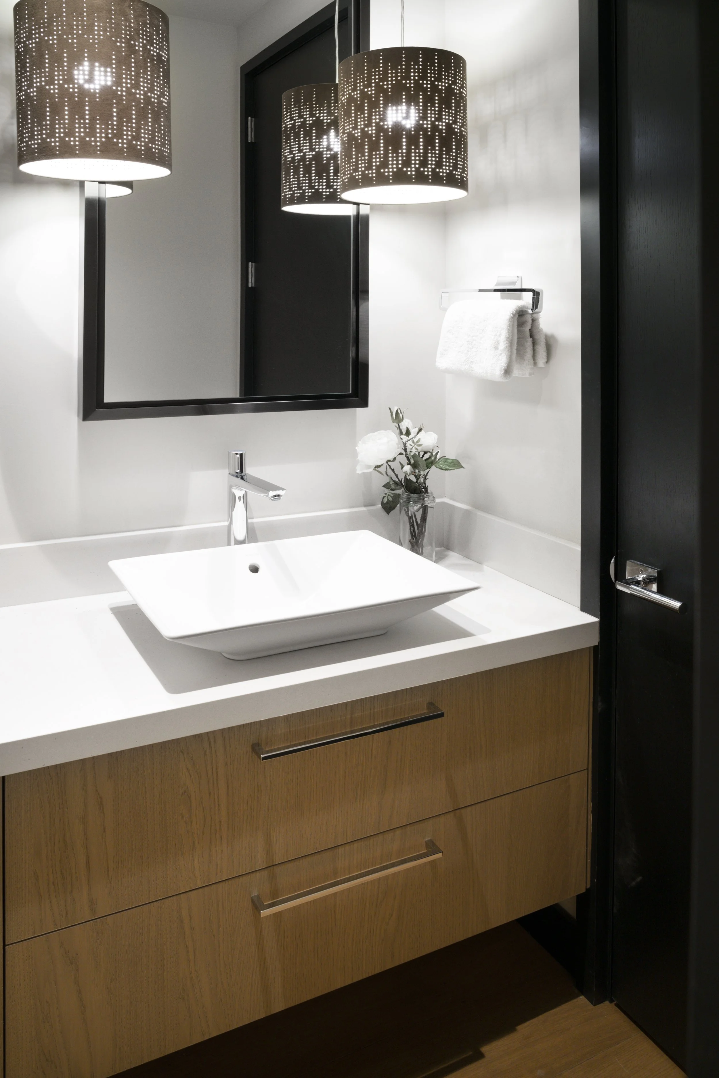 Modern bathroom sink with a white vessel basin, wooden cabinet, large wall mirror, black framed door, decorative flower vase, and two hanging pendant lights with perforated black shades.