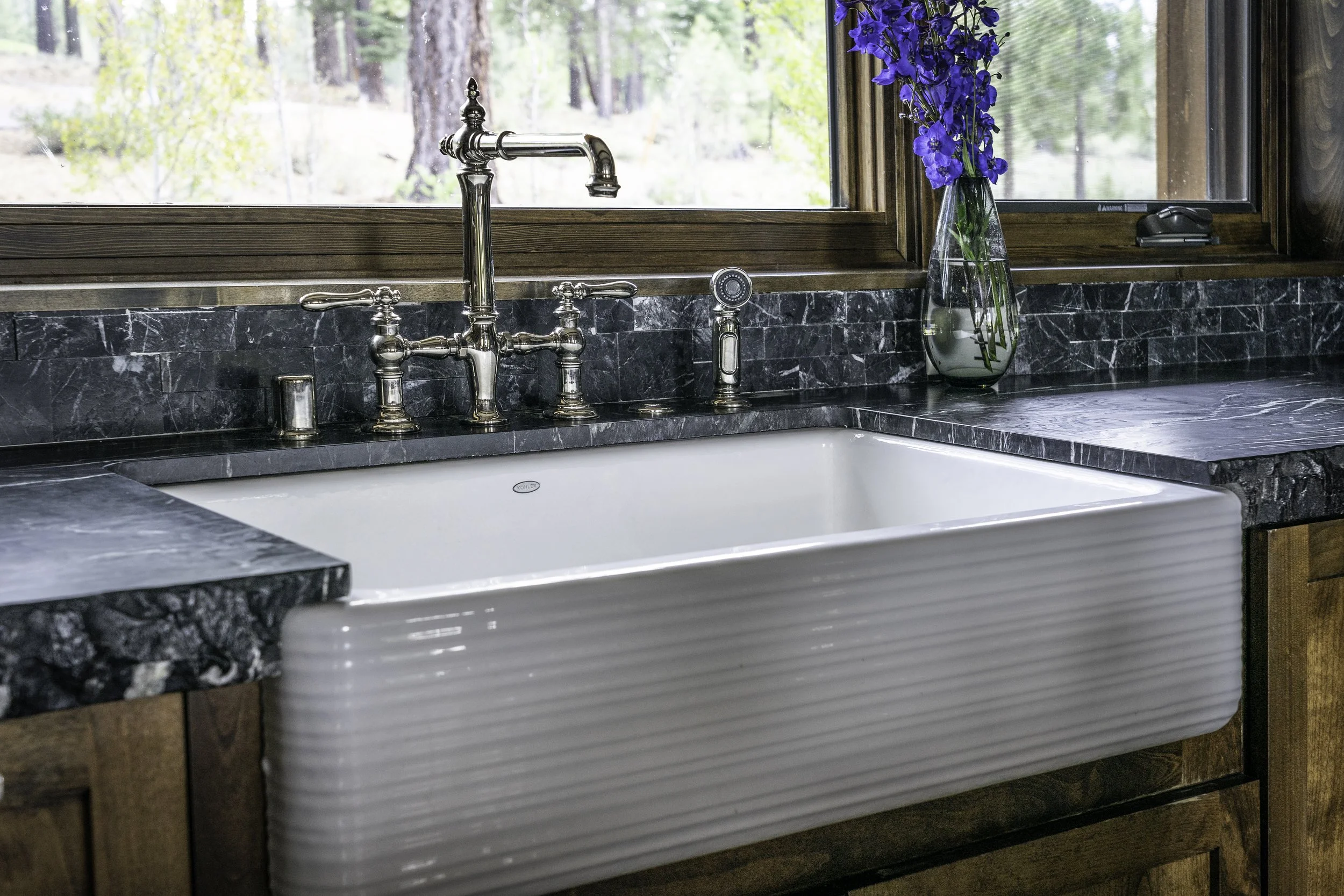 Kitchen sink with a black marble countertop, vintage-style silver faucet, and a window with a view of trees. A vase with purple flowers is on the right side of the sink.
