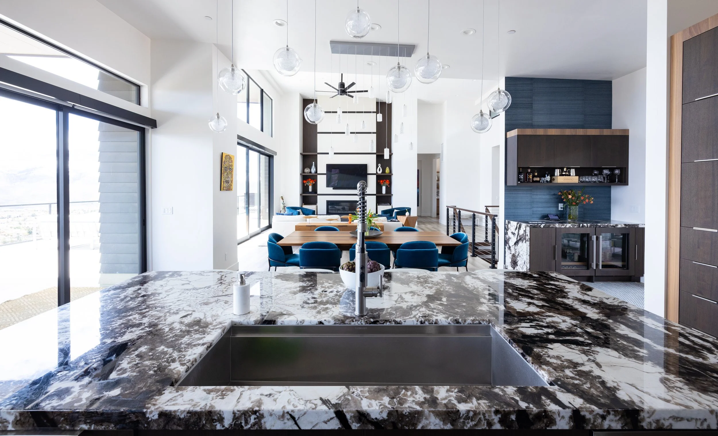Modern open-concept kitchen and dining area with marble countertop, blue chairs around a wooden table, large windows, and a living room with a fireplace and TV in the background.
