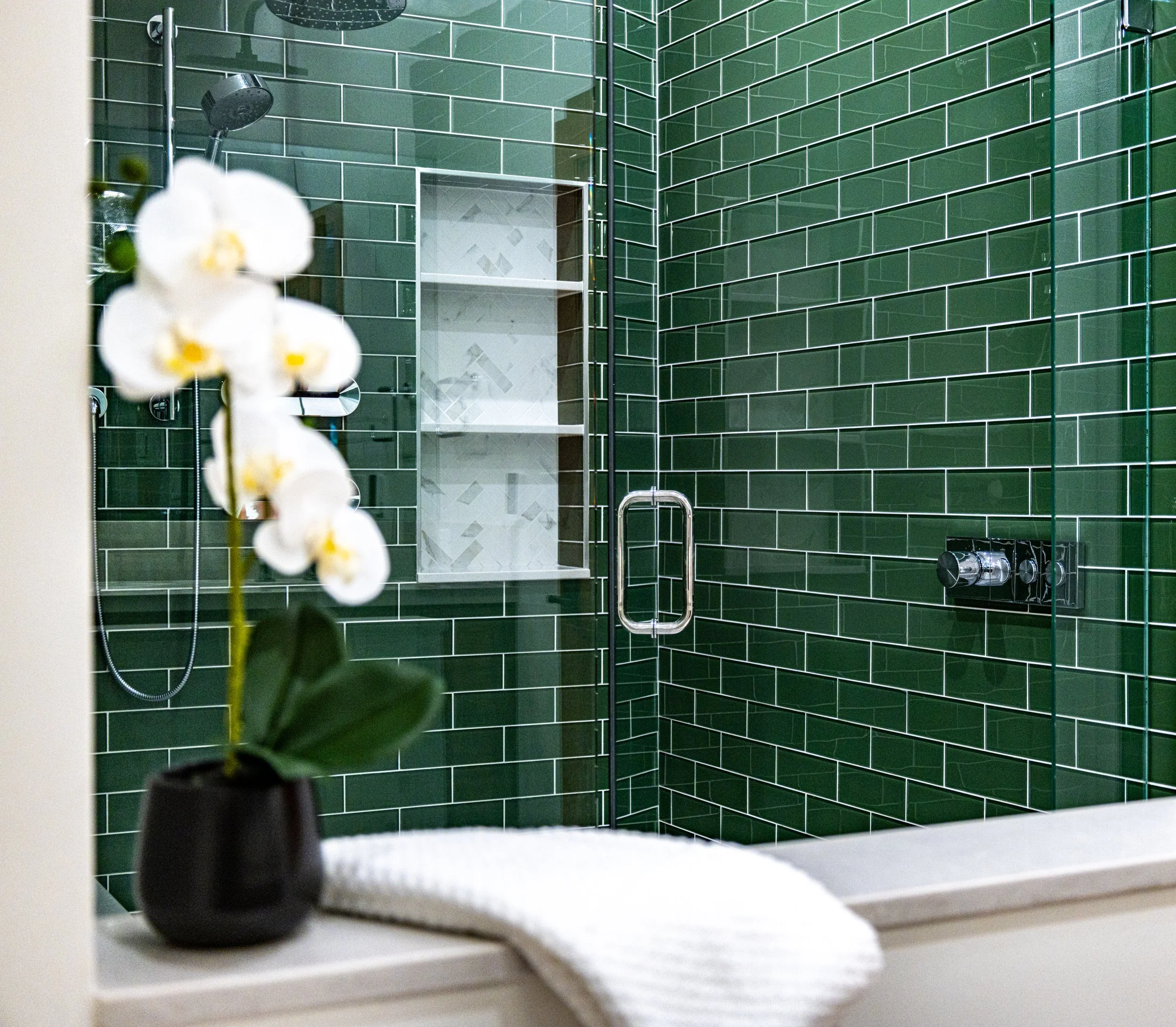 Green tiled shower with a glass door, built-in shelf, and a walk-in shower head, seen through a bathroom window with a potted orchid and a white towel in the foreground.