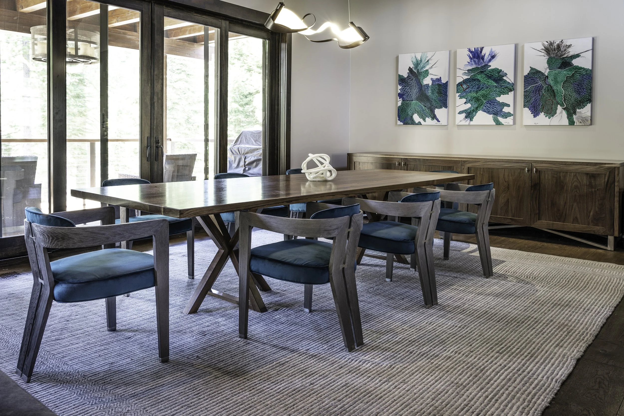 A dining room with a wooden table and blue cushioned chairs, a light gray rug, artwork of blue and green leaves on the wall, and large glass sliding doors leading to a balcony with outdoor furniture.