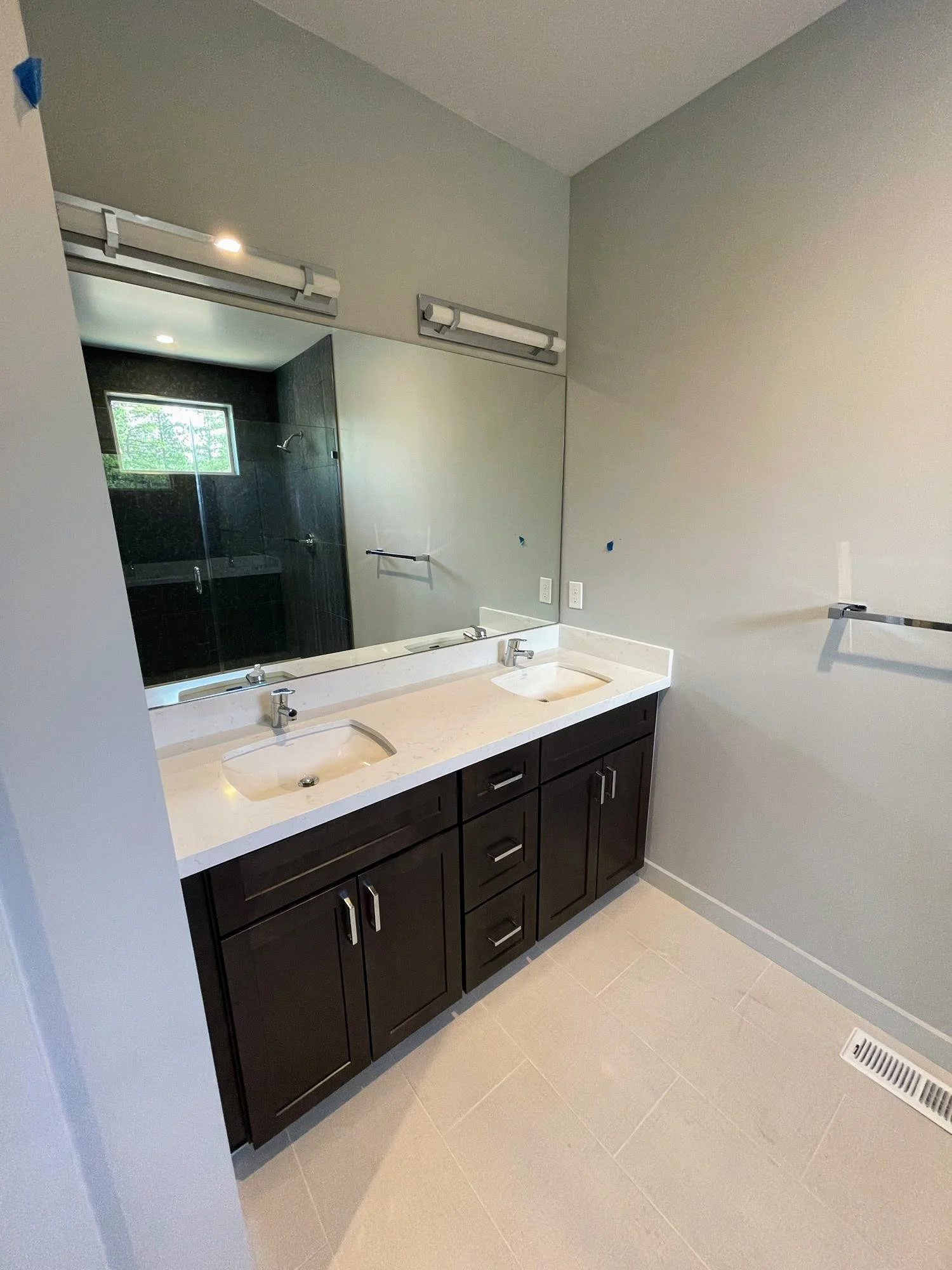 Bathroom with double vanity, dark wood cabinets, white countertop, two sinks, large mirror, and a walk-in shower visible in the reflection.