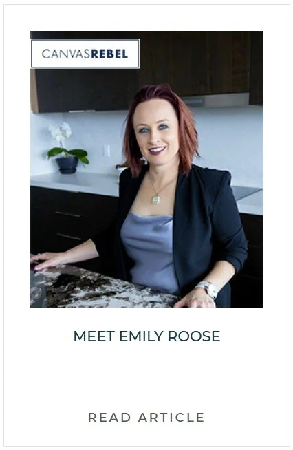 A woman with red hair, wearing a black blazer and a gray top, sitting in a modern kitchen with dark cabinets and a marble countertop, smiling at the camera.