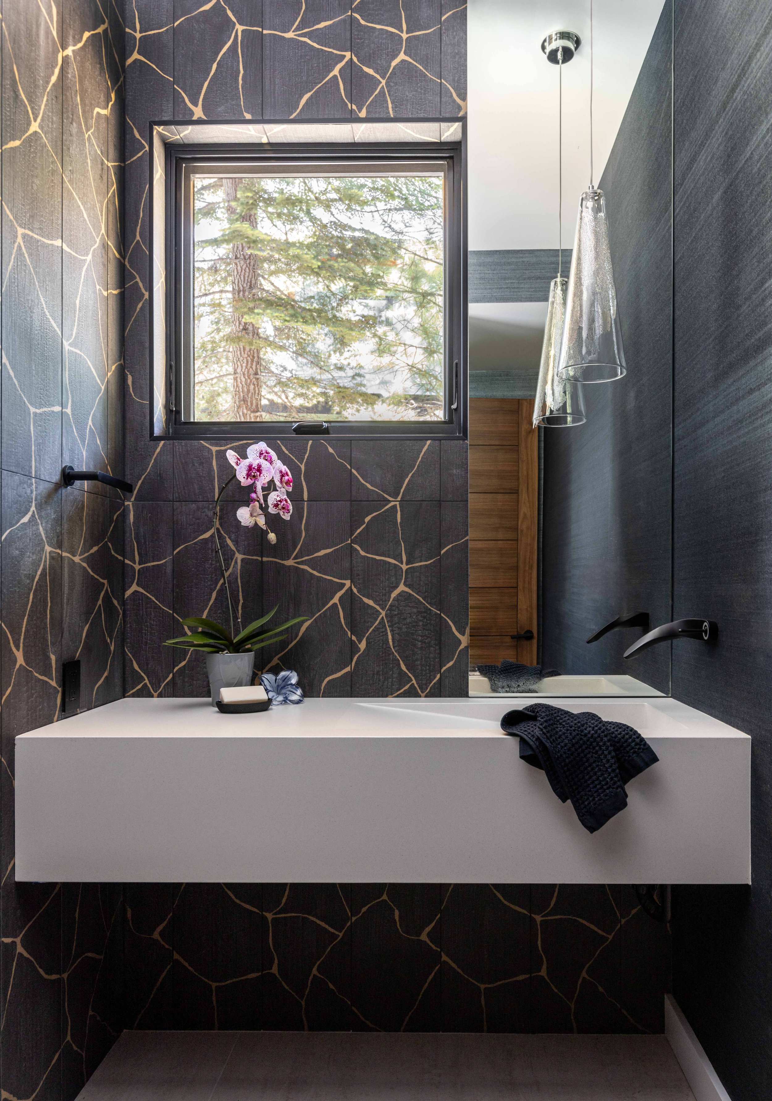 Modern bathroom with dark, patterned tile walls, a rectangular white sink, black fixtures, a window with a view of trees, and hanging glass pendant lights.