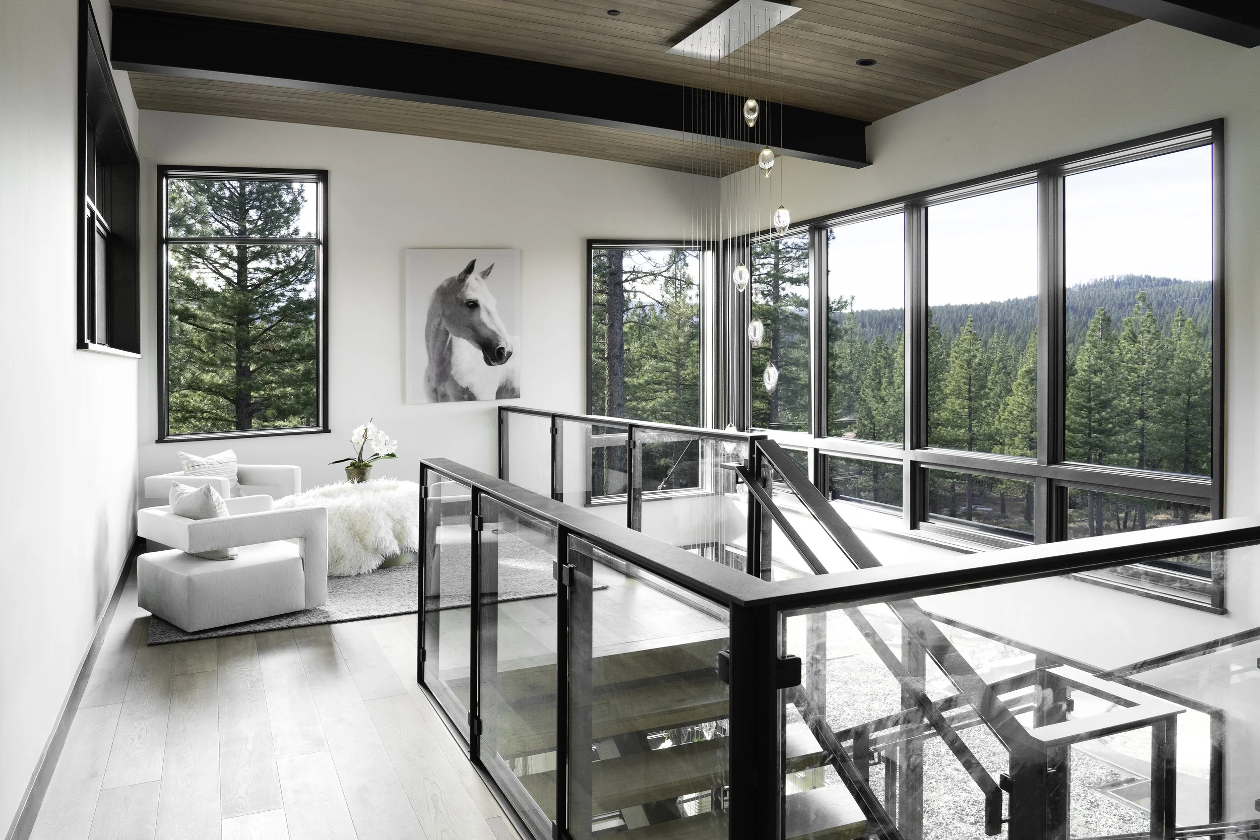 Modern interior with large windows overlooking a forest, white chairs with pillows, and a white horse portrait.