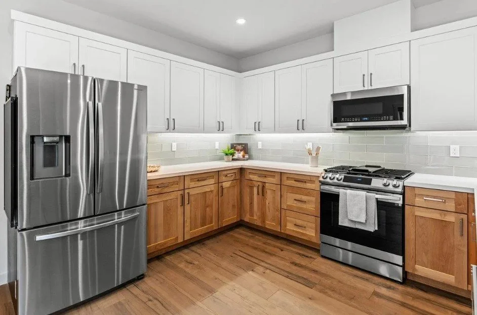 Modern kitchen with stainless steel refrigerator, gas stove, microwave, and wooden cabinets with white countertops and light gray tile backsplash.