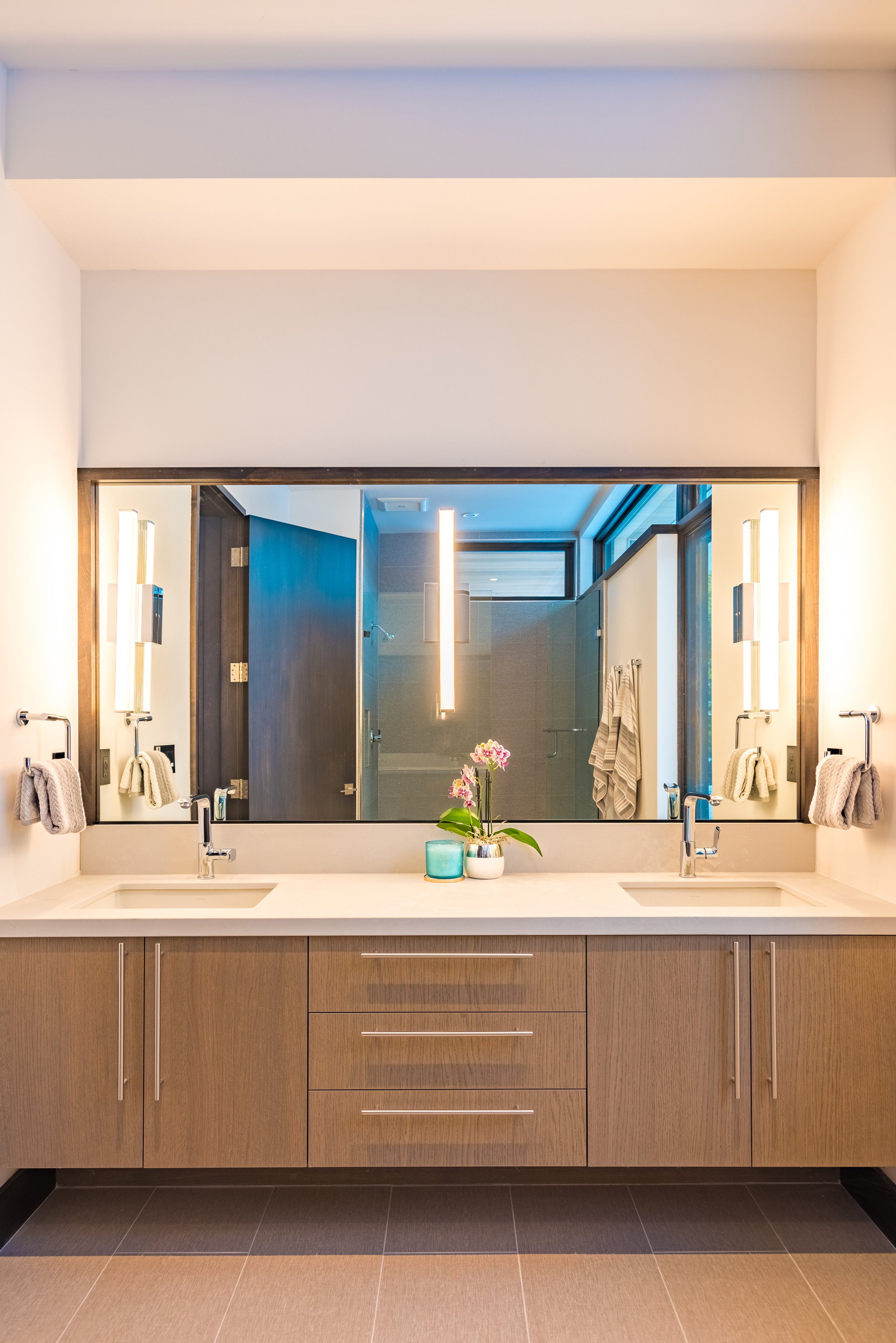 Modern bathroom with double sinks, large mirror, and wooden vanity, decorated with a potted orchid and candles, with a shower visible in the background.