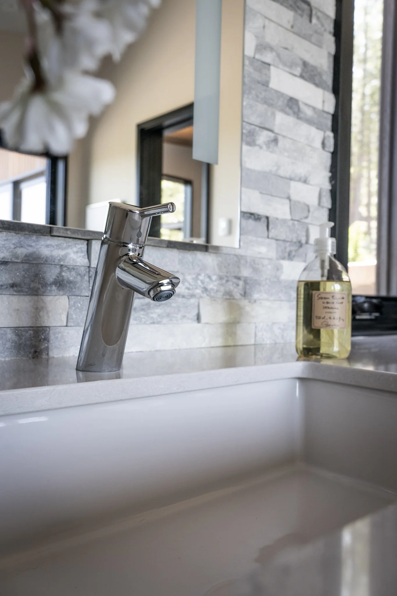 Close-up of a modern kitchen sink with a chrome faucet, surrounded by a stone backsplash and a bottle of soap on the countertop.