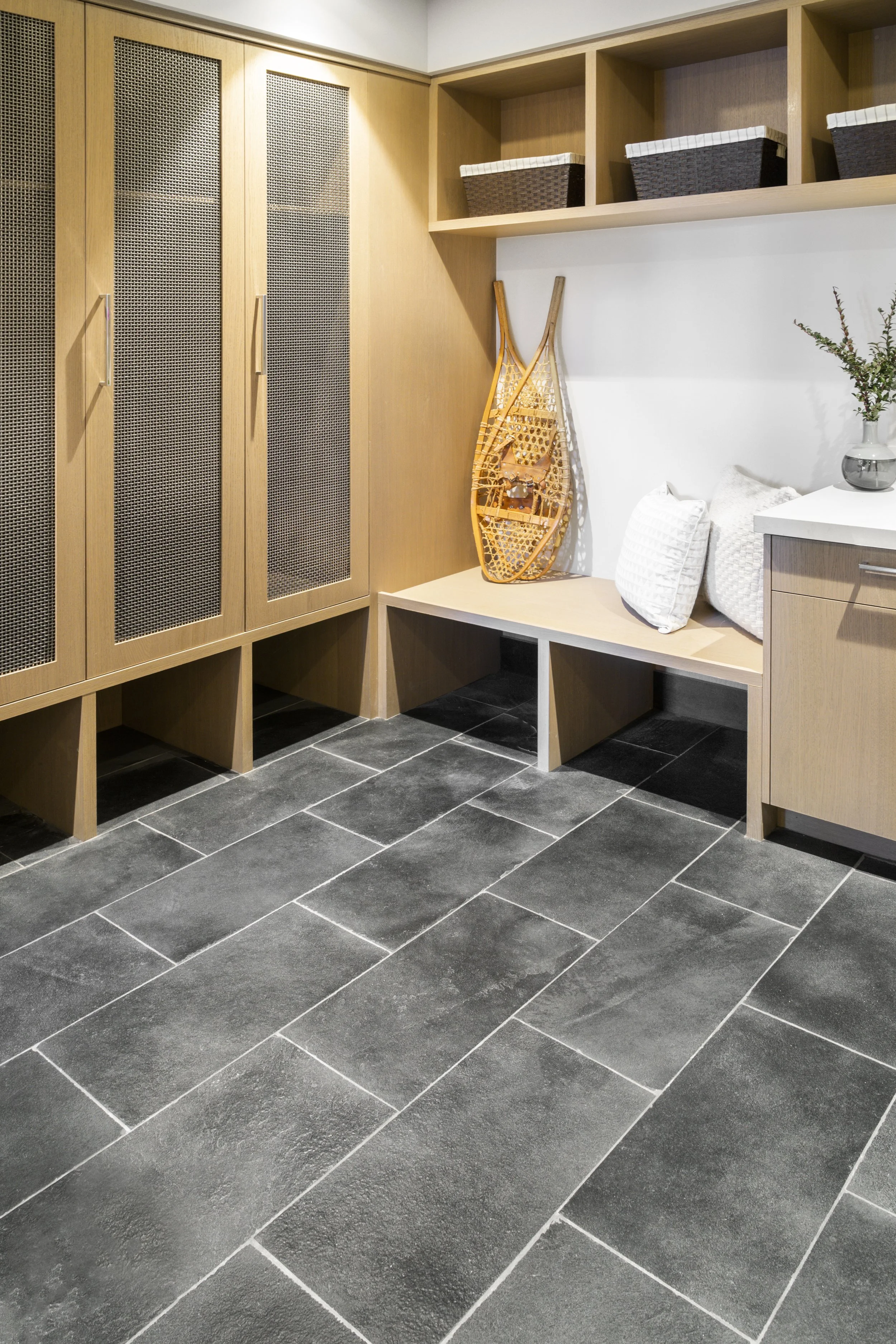 A modern entryway with wooden cabinets, woven baskets, a decorative sled, pillows, and a potted plant on a white cabinet, with black tiled flooring.