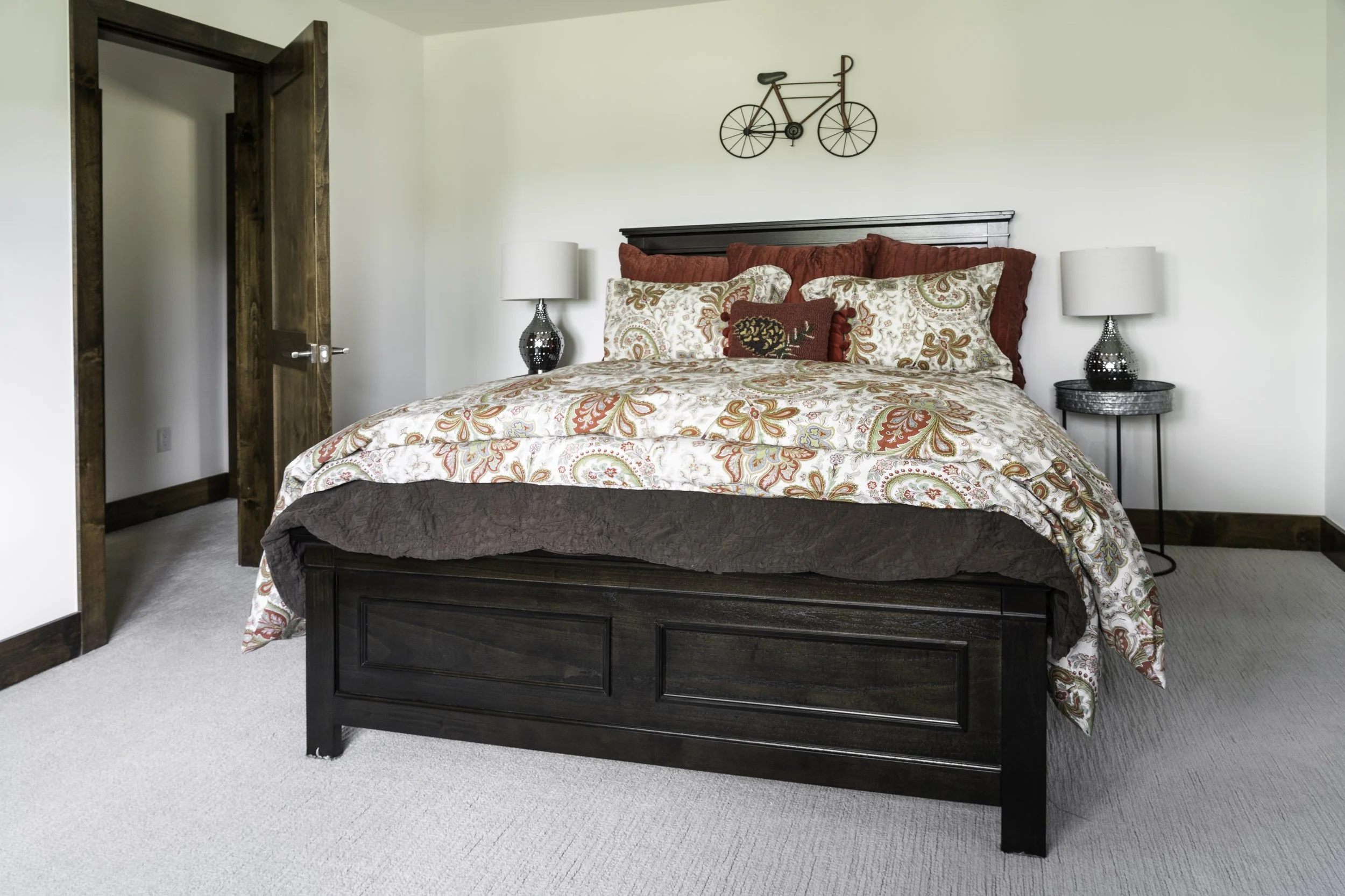 A bedroom with a large bed with multicolored floral and paisley bedding, red and patterned pillows, a dark wood headboard, and matching dark wood frame. Two white table lamps with black bases are on black side tables. There is a bicycle-shaped wall d