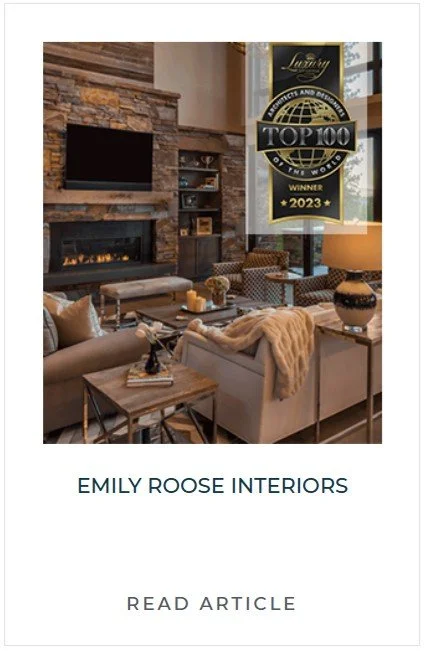 Interior of a modern living room with a stone fireplace, flat-screen TV, comfortable seating, and decorative lamps. A banner with a "Top 100 of the World" award from 2023 hangs in the room.