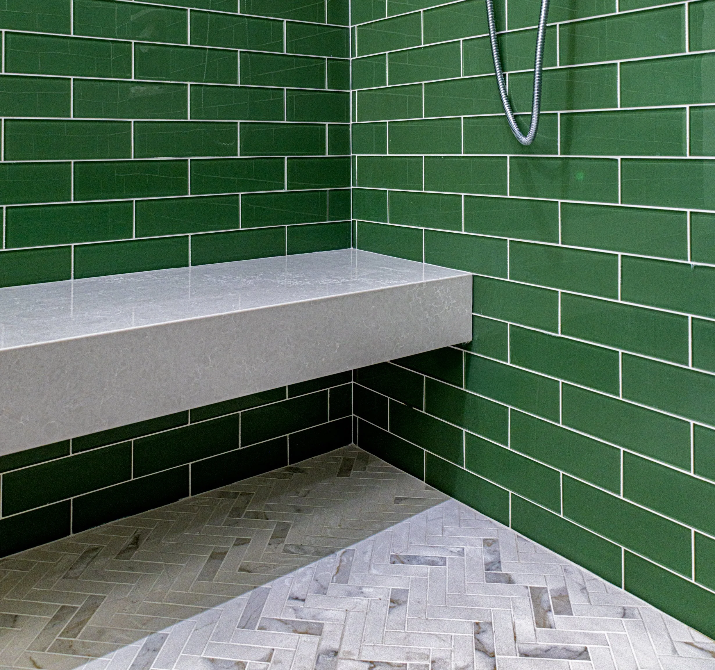 A corner of a shower with green subway tiles on the walls, a gray concrete shelf, and a patterned gray tile floor.