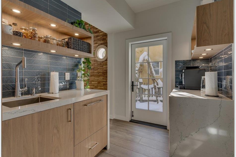 Modern kitchen with wooden cabinets, black tile backsplash, open shelves with jars, and a snowy outdoor scene visible through a glass door.