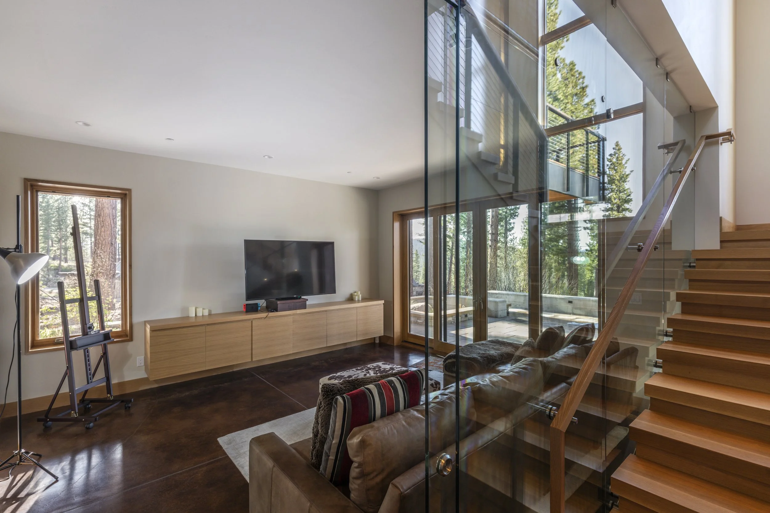 Modern living room with large glass windows and sliding door view of a wooded outdoor patio, staircase with wooden steps and glass railing, a flat-screen TV on a minimalist wooden console, a leather couch with striped pillows, and a tall floor lamp.