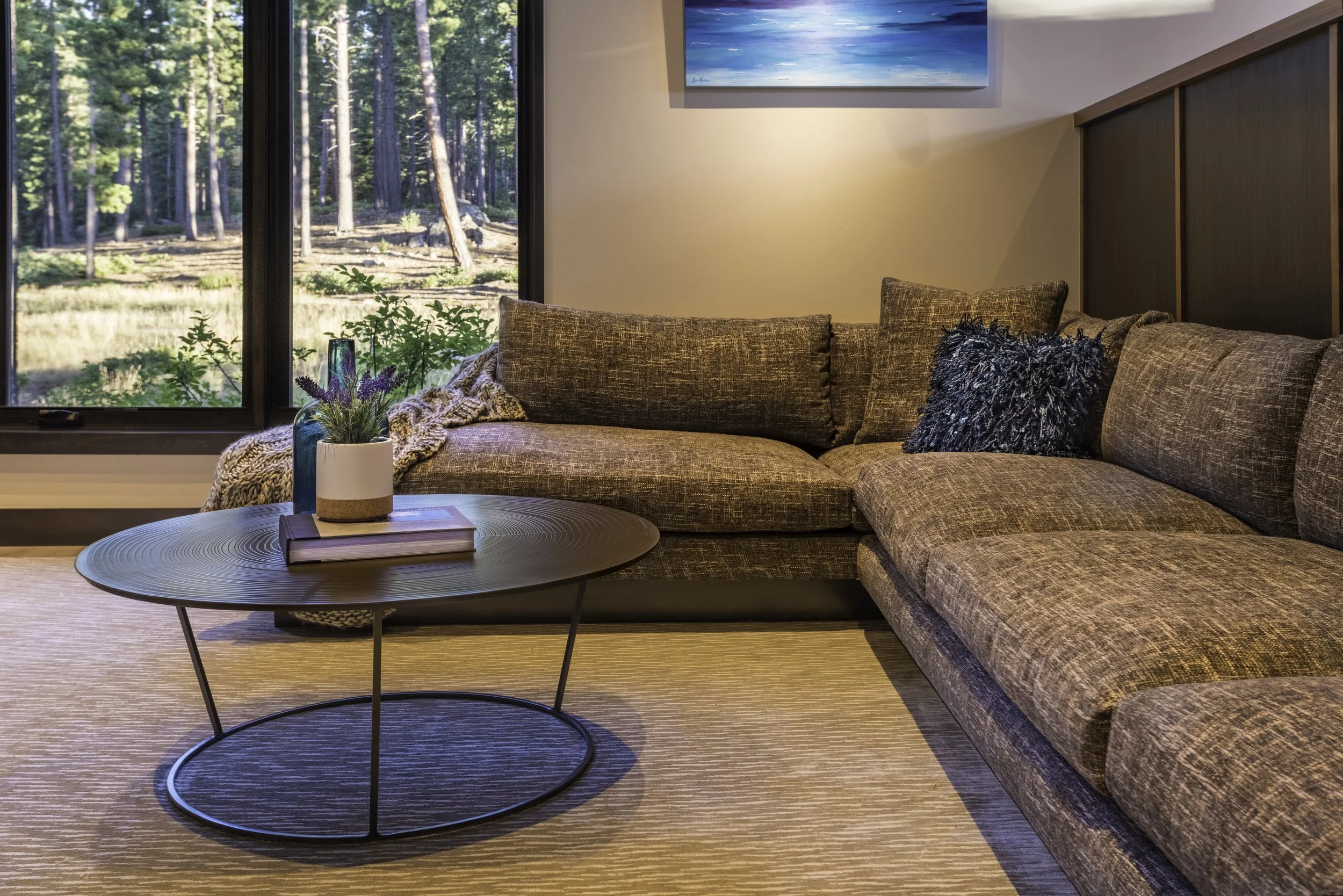 Living room with a brown sectional sofa, a round black coffee table with a plant and books, large window showing a forest outside, and a painting on the wall.