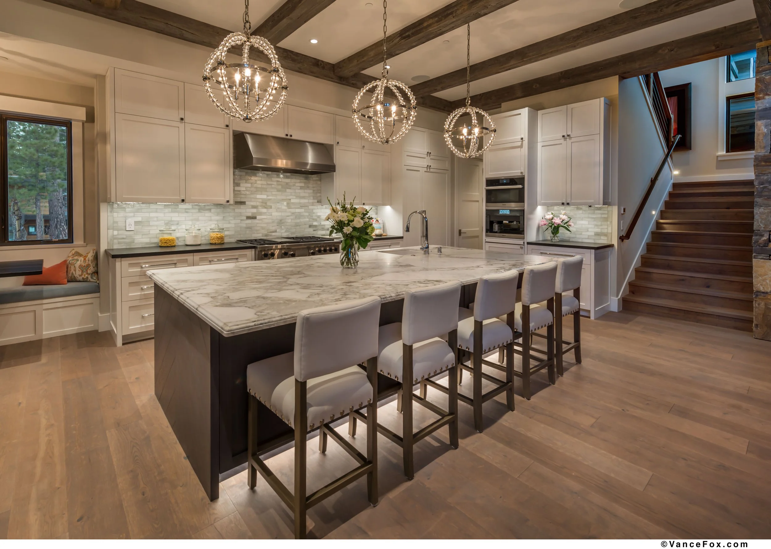 Modern kitchen with a large marble island, white cabinets, pendant chandeliers, hardwood floors, and a staircase to the right.