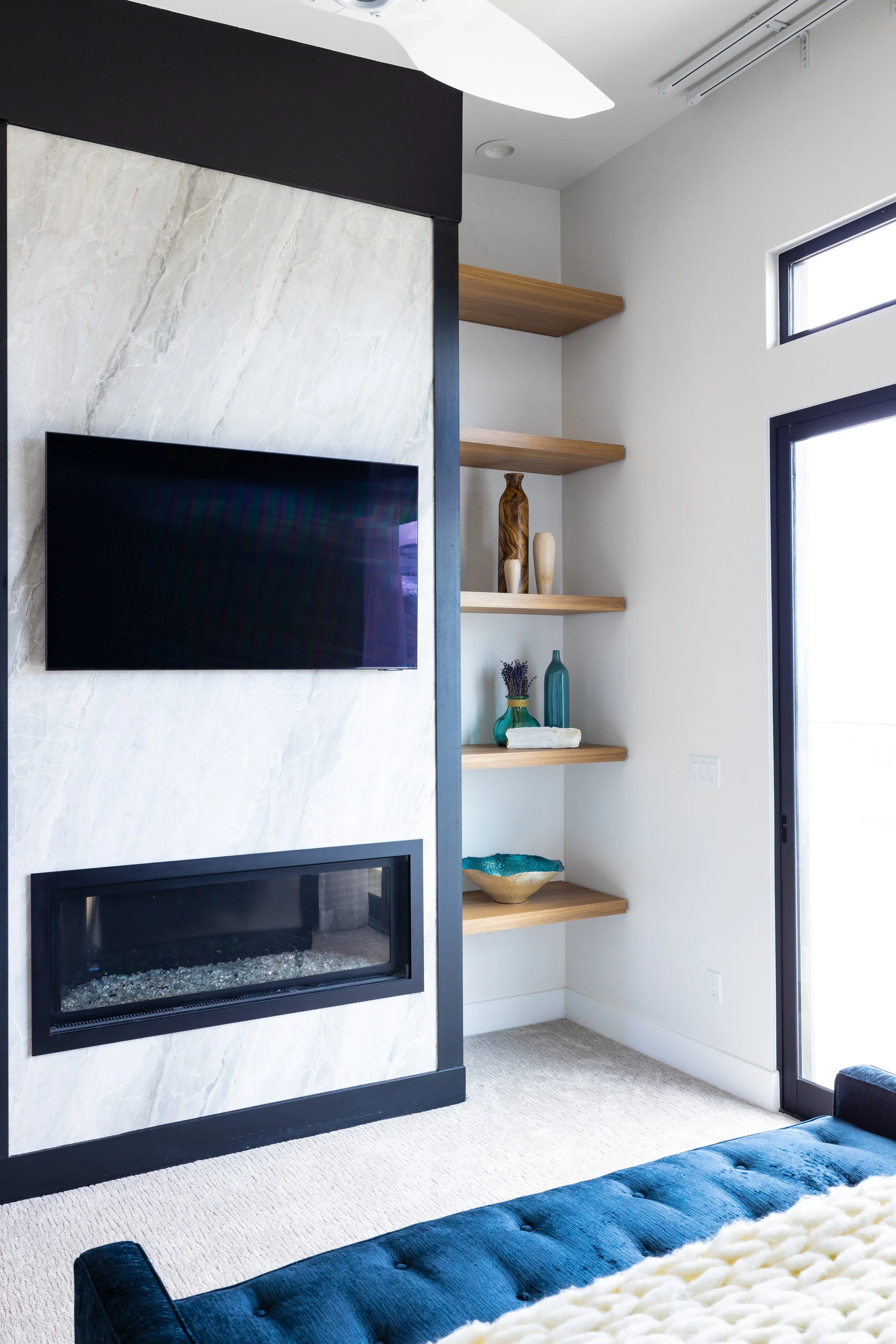 Modern living room with a flat-screen TV, a fireplace, and floating shelves with decorative vases and bowls near a large window.