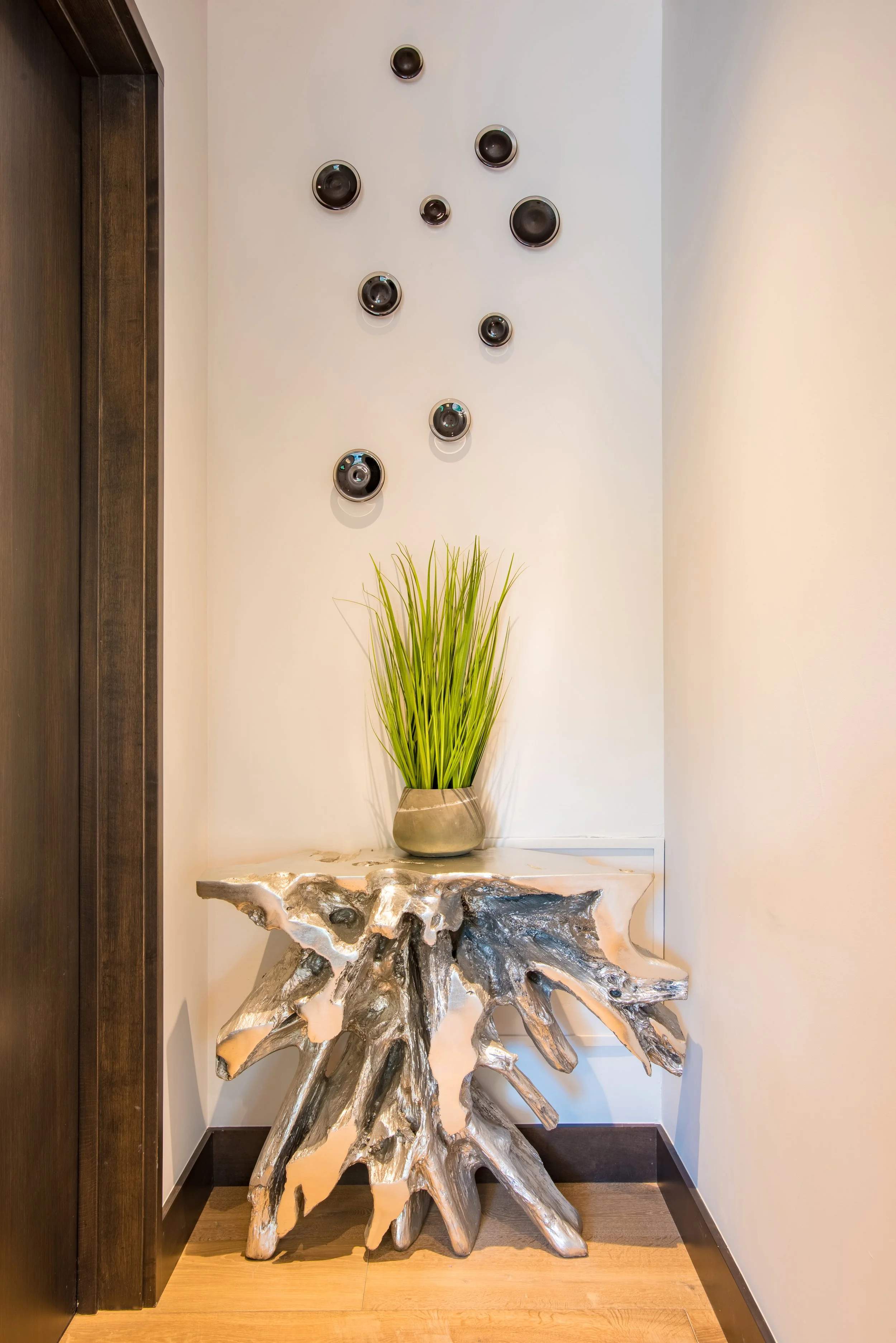 Decorative wall with black speakers, a ceramic vase with green grass, and a sculptural wooden table.