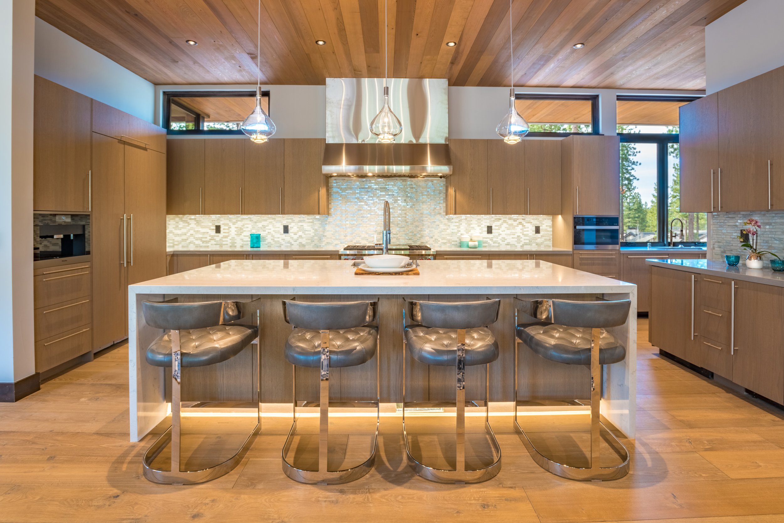 Modern kitchen with a large white island counter, four metallic bar stools, wood cabinets, a backsplash, and large windows providing natural light.