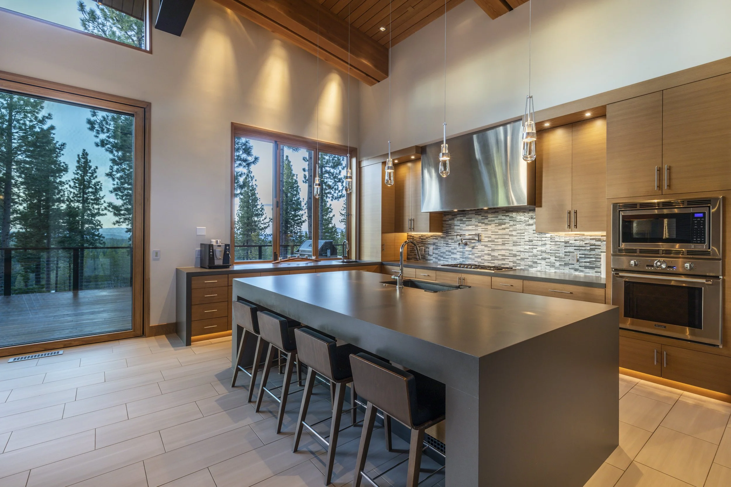 Modern kitchen with large island, four black bar stools, stainless steel appliances, and large windows showing trees outside.