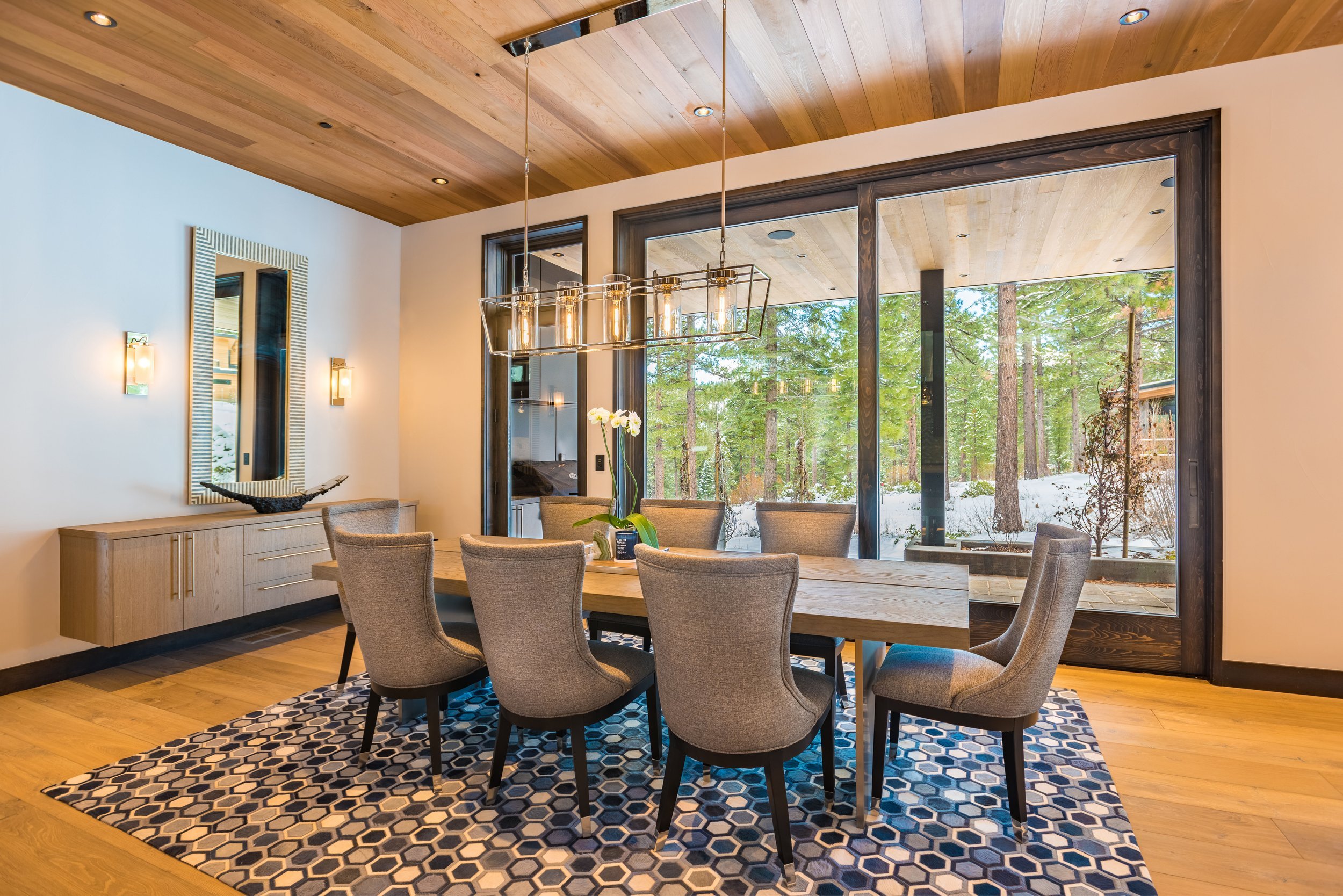 Modern dining room with a large wooden table surrounded by eight beige upholstered chairs. A geometric-patterned rug is underneath the table. Large glass doors reveal a snowy forest outdoors. Wooden ceiling with recessed lighting, wall-mounted lamps,