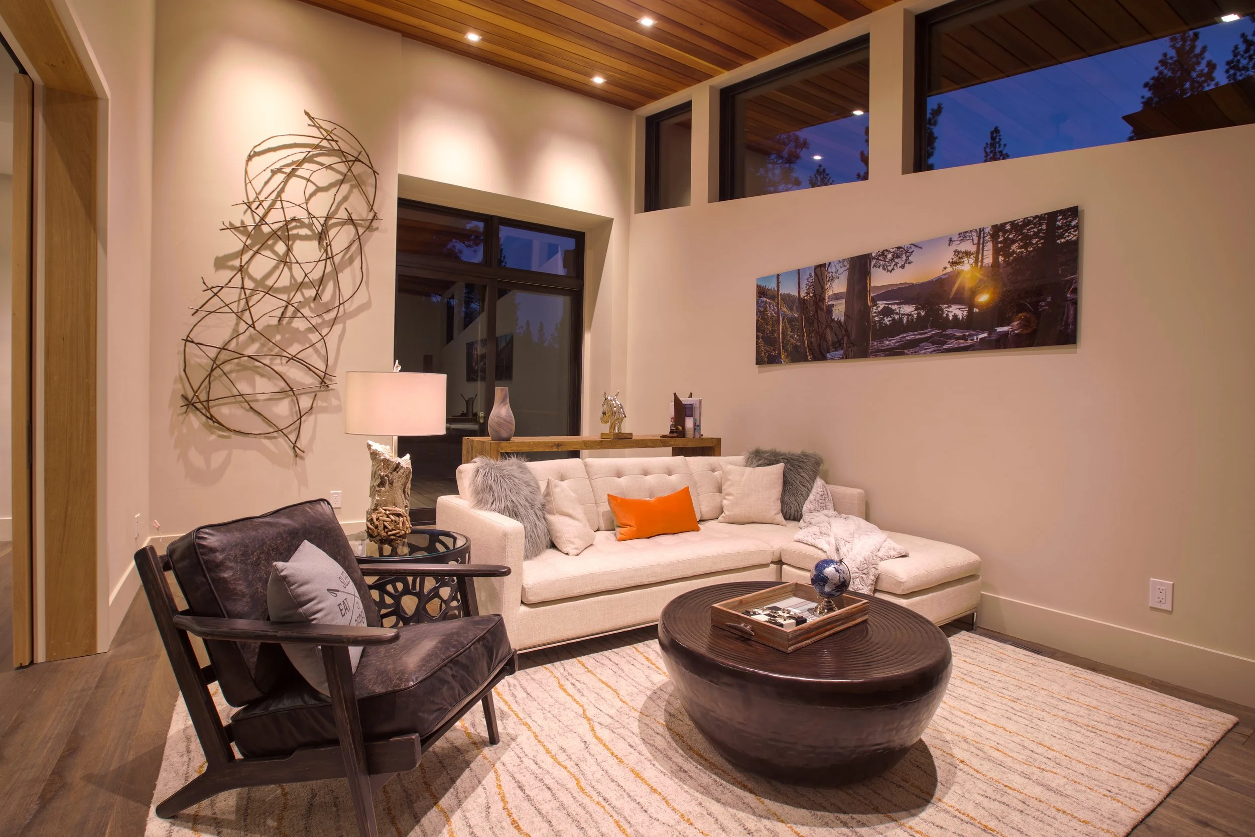 Living room with cream sofa, black armchair, oval wooden coffee table, artwork on wall, windows showing outdoor scenery at dusk, decorative items, and warm lighting.