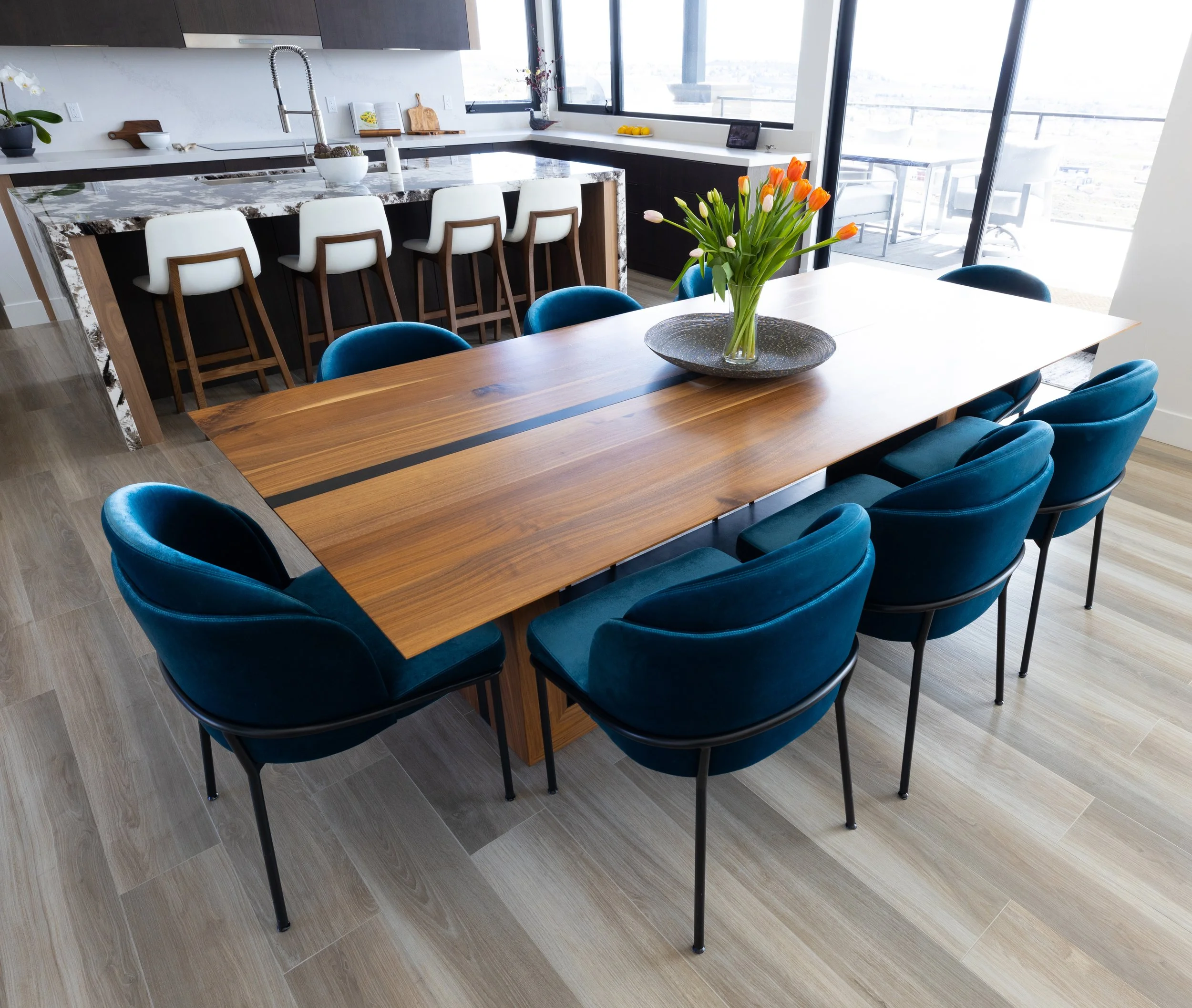 Modern dining room with a large wooden table surrounded by ten velvet upholstered chairs in dark teal. A vase with orange and white tulips is centered on the table. In the background, an open kitchen with a marble island, white barstools, and large w