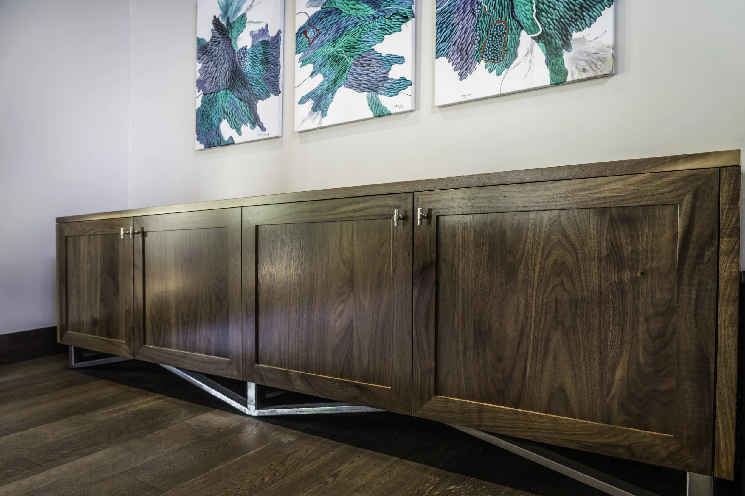 A wooden cabinet with metal legs in a room with gray walls and wooden floor, decorated with three framed artwork pieces featuring blue and green abstract designs of leaves or feathers.