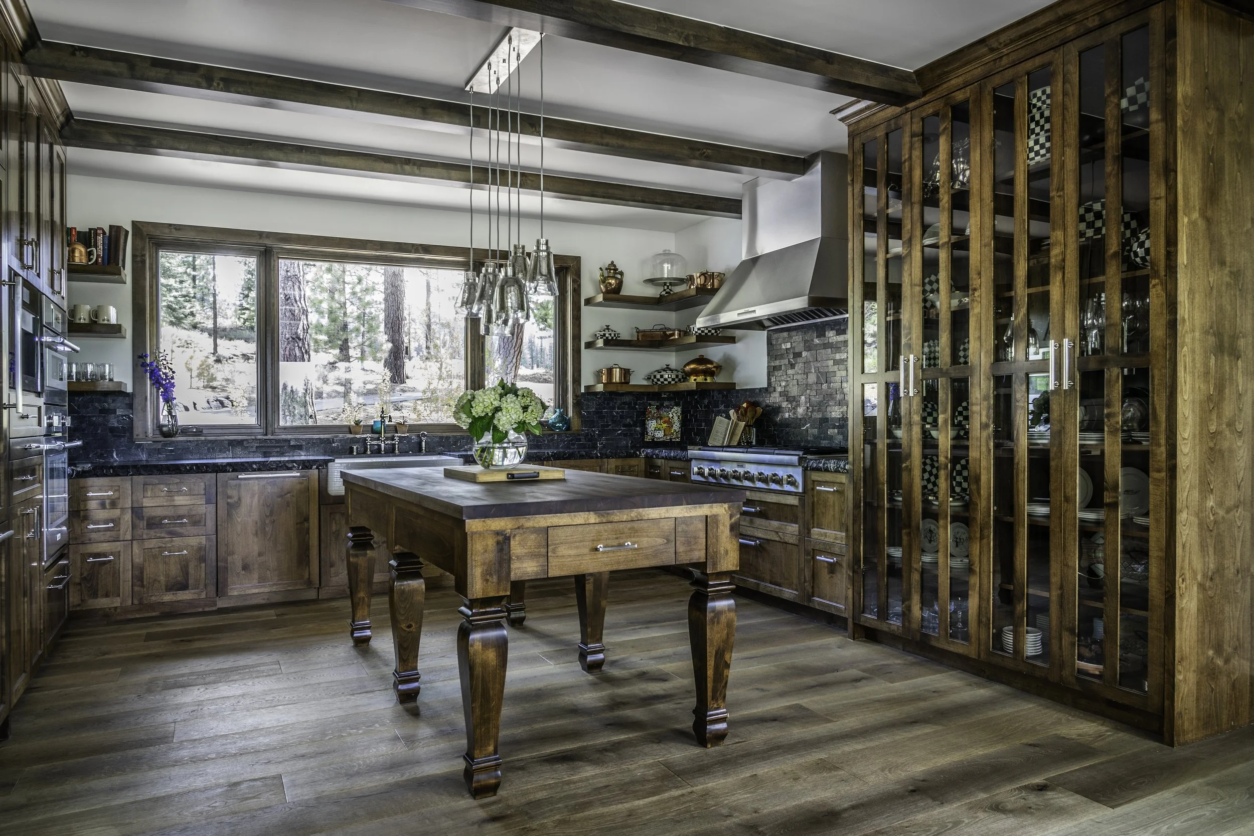 A cozy kitchen with wooden cabinetry, a central wooden table with a flower vase, large window overlooking trees, modern stove and hood, hanging pendant lights, open shelves, and a tall wooden glass-fronted cabinet filled with dishes and glassware.