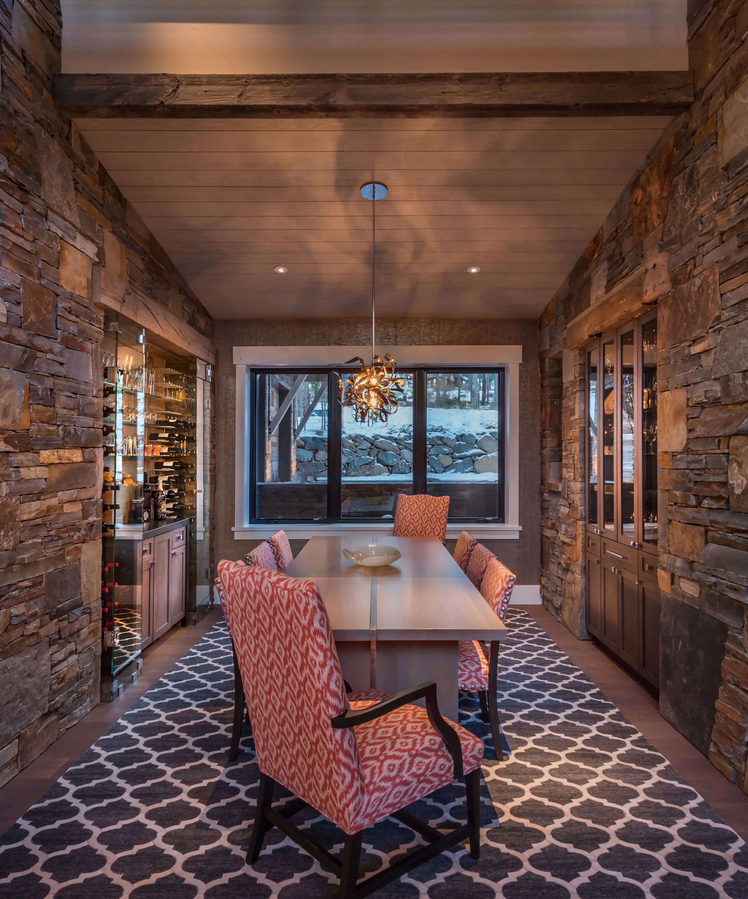A cozy dining room with stone walls, a long wooden table, and six patterned upholstered chairs. A contemporary chandelier hangs above the table. Large windows look out onto a snowy landscape with rocks and trees.