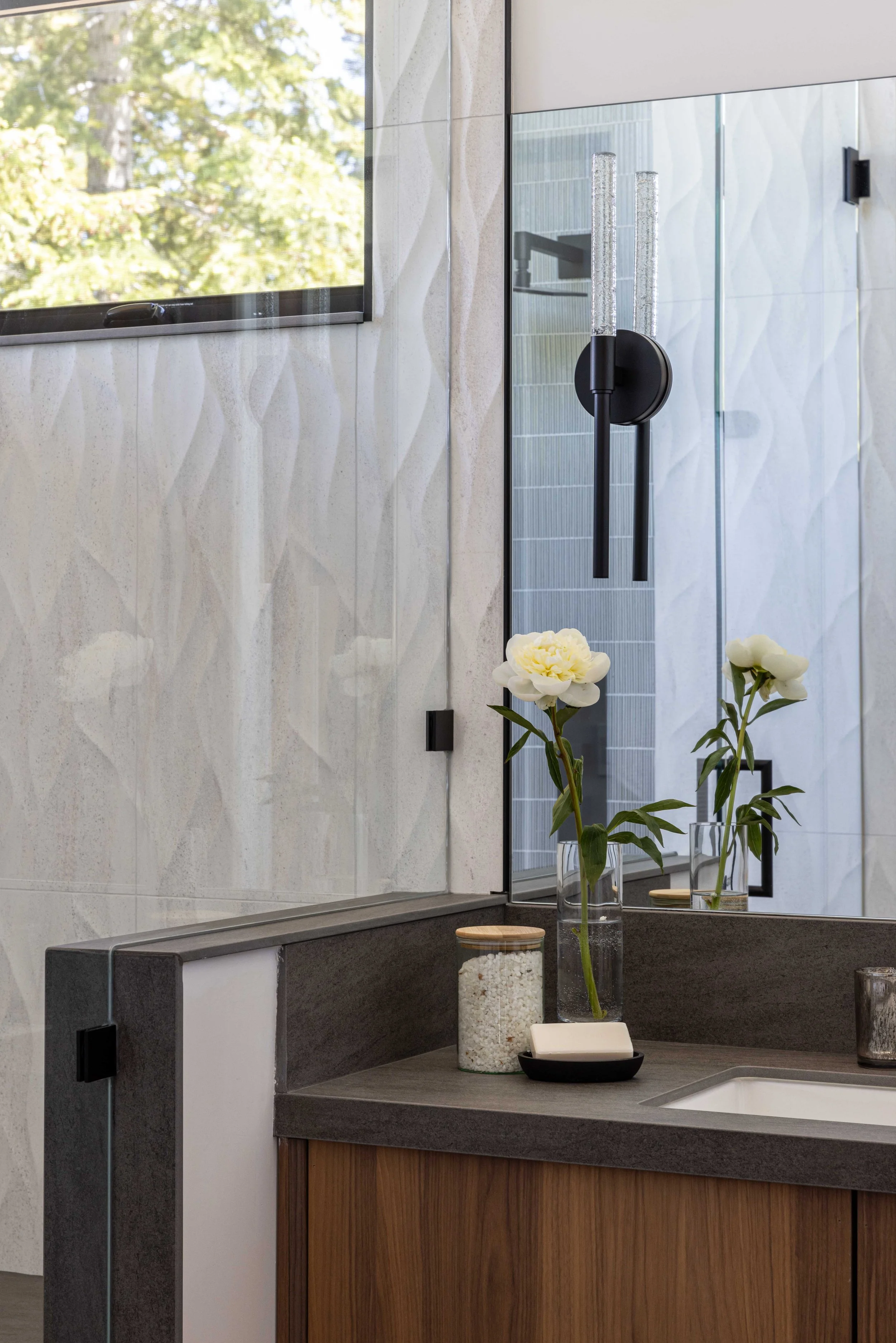 A bathroom with a large mirror, a soap dish, a jar of bath salts, and a vase with white flowers on the countertop. There is a window reflecting trees outside.
