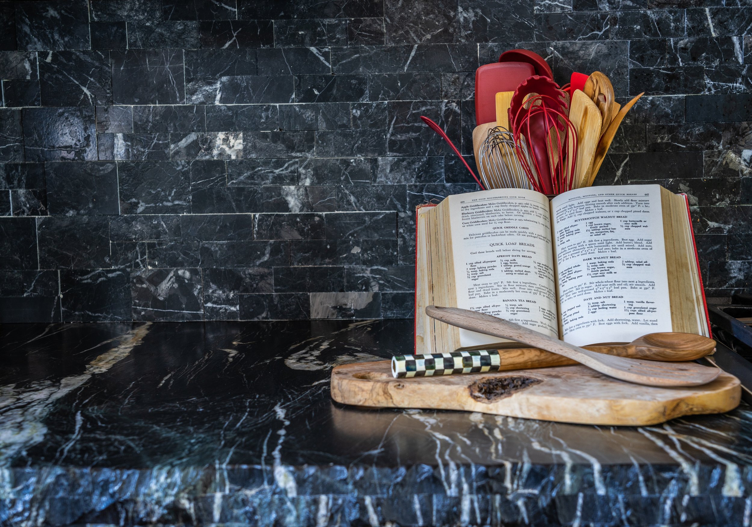 Kitchen countertop with an open recipe book, cutting board, and various kitchen utensils, including spoons and whisks in a container, against a dark marble backsplash.