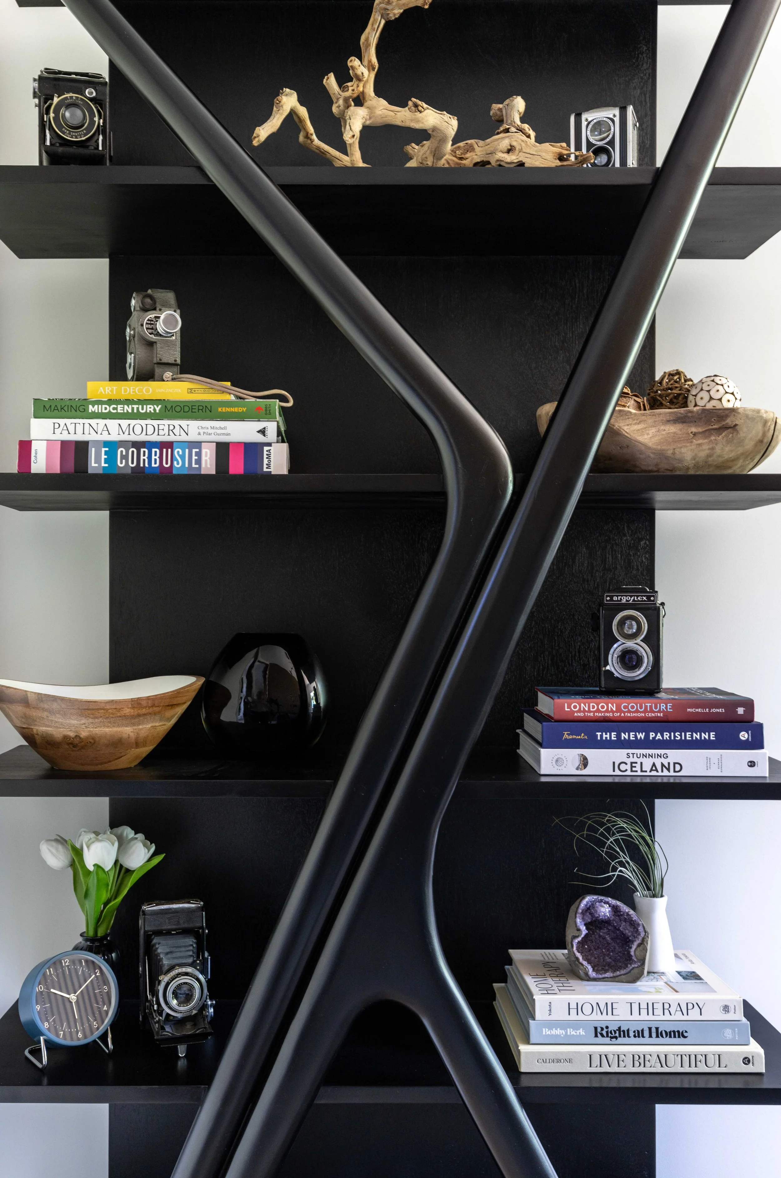 Black shelving unit holding decorative items, books, vintage cameras, vases, a clock, and an amethyst geode, with a modern black metal sculpture in front.