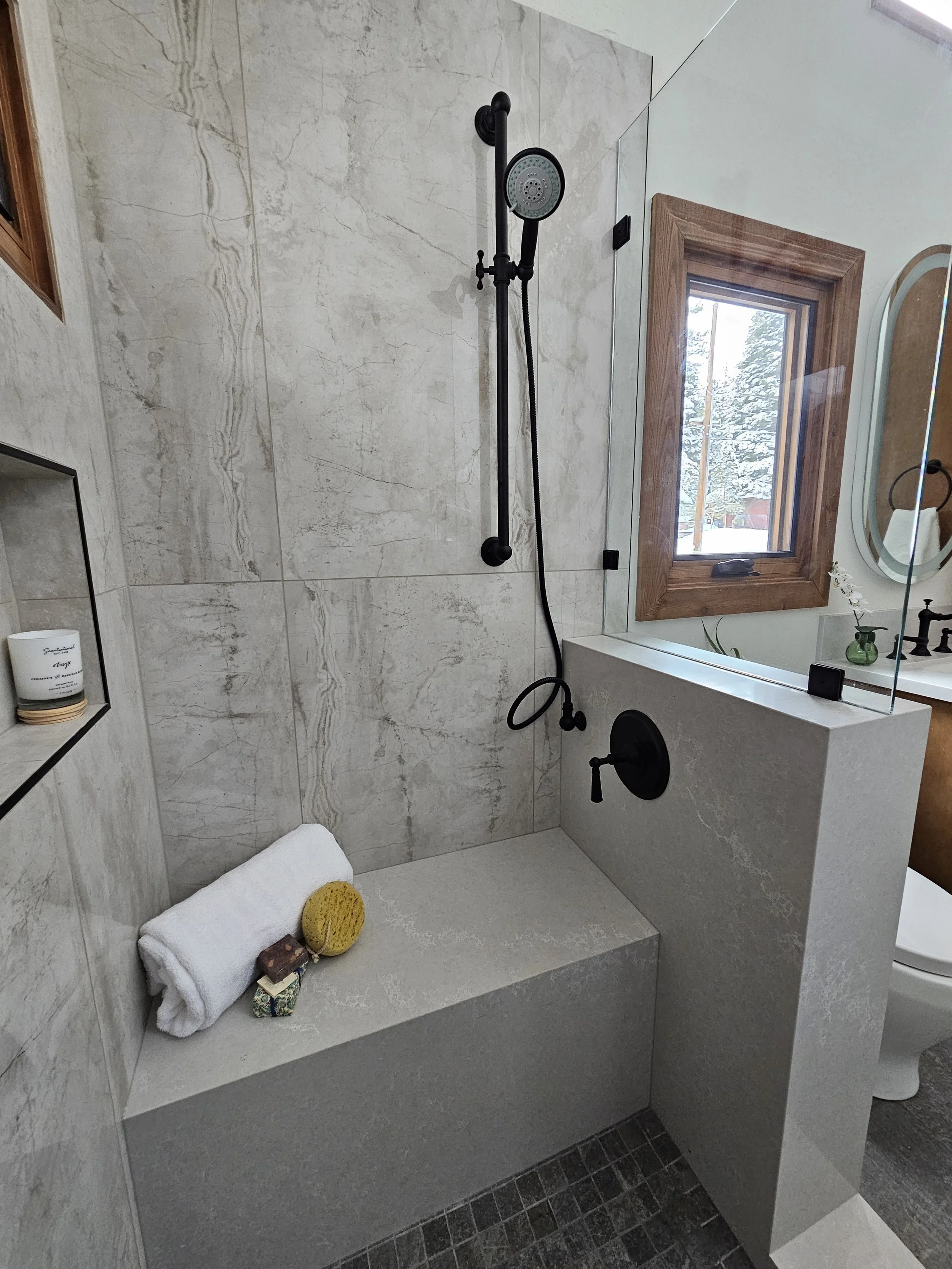 A modern bathroom shower area with large marble tiles, a black shower head and fixtures, a wooden-framed window with outside snow and trees, and a built-in niche with bath products. There are towels and a sponge on a bench, and a partial view of a to