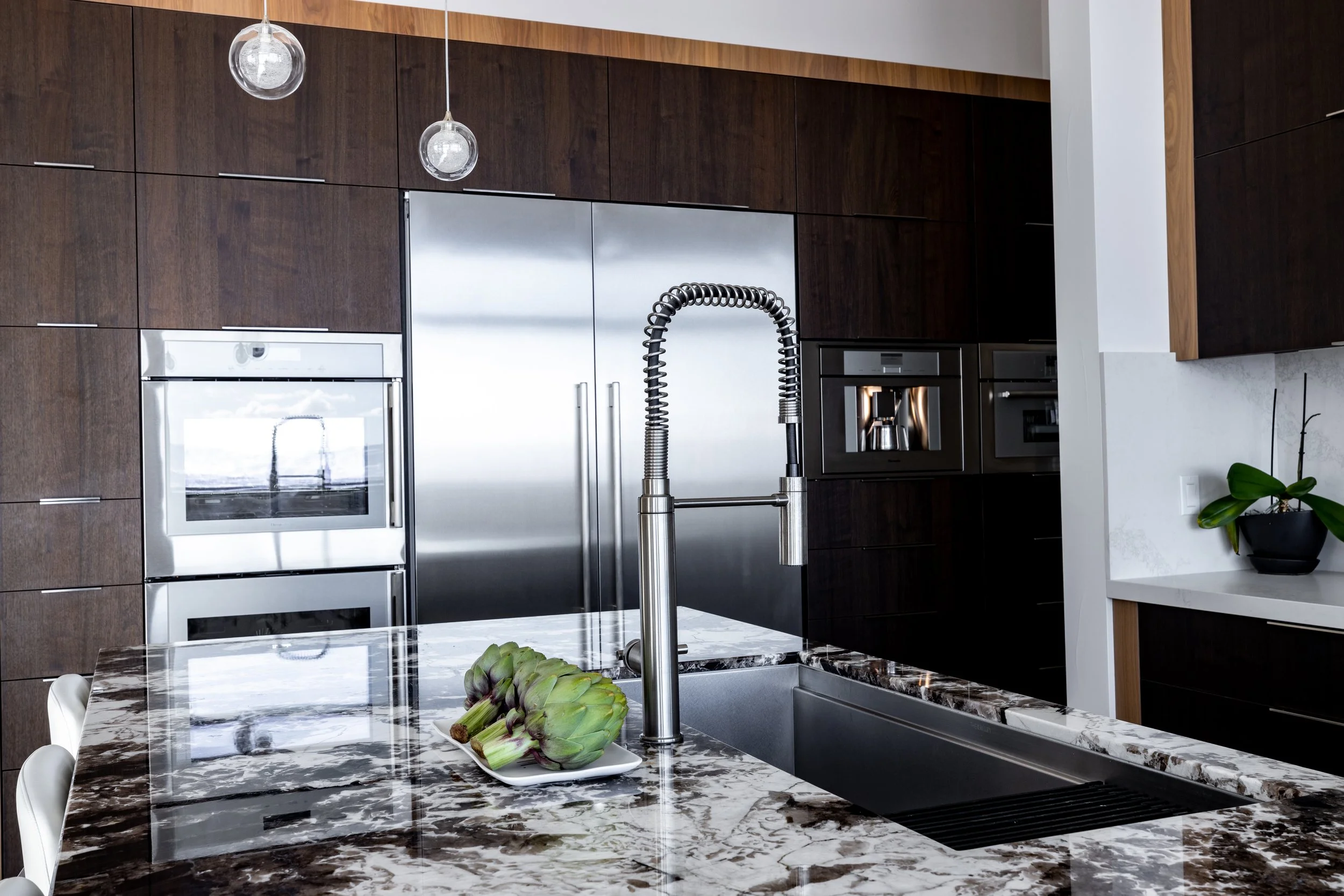 Warm modern kitchen with stainless steel appliances, dark wood cabinets, marble countertop, and a sink with a high-arc faucet. There are two artichokes on a white tray on the countertop, and a potted plant on a white ledge to the right.